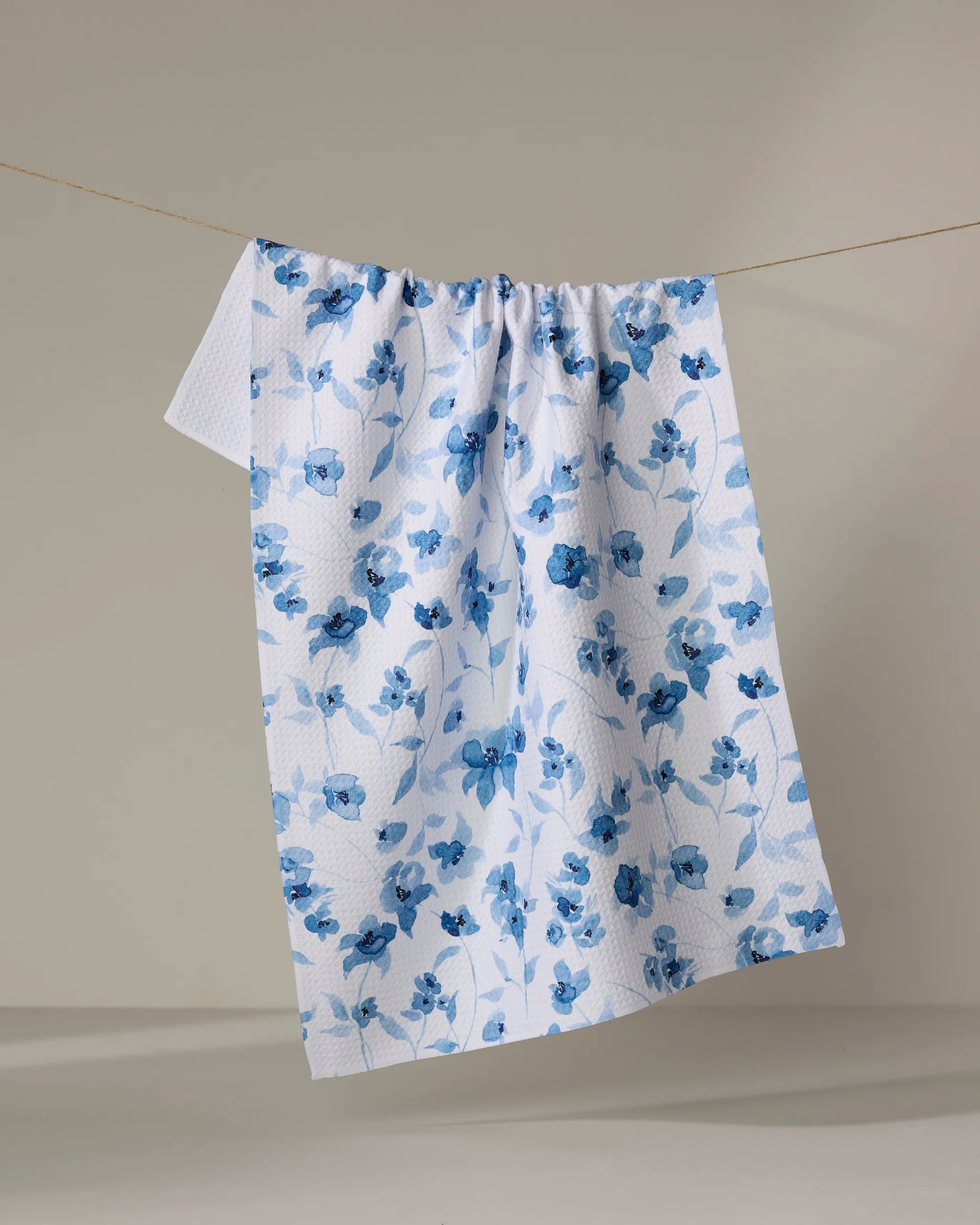 The "Blue Floral" kitchen tea towel by Aubrey Fairchild, featuring a blue floral pattern and made from recycled materials, hangs on a clothesline against a neutral background.
