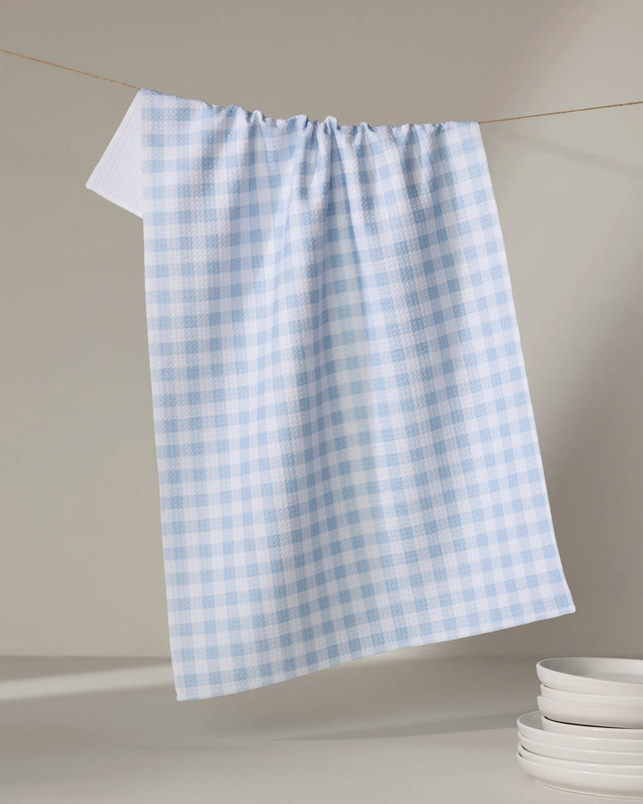 The Aubrey Fairchild Blue Gingham tea towel, crafted from recycled materials, hangs on a string beside stacked white plates on a light surface.