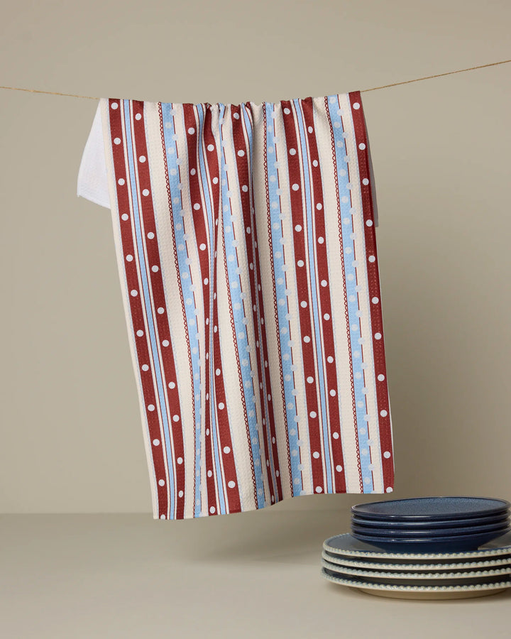 The Geometry Blue Skies Stripes kitchen towel, crafted from recycled materials, hangs beside a stack of blue and white striped plates on a neutral background.