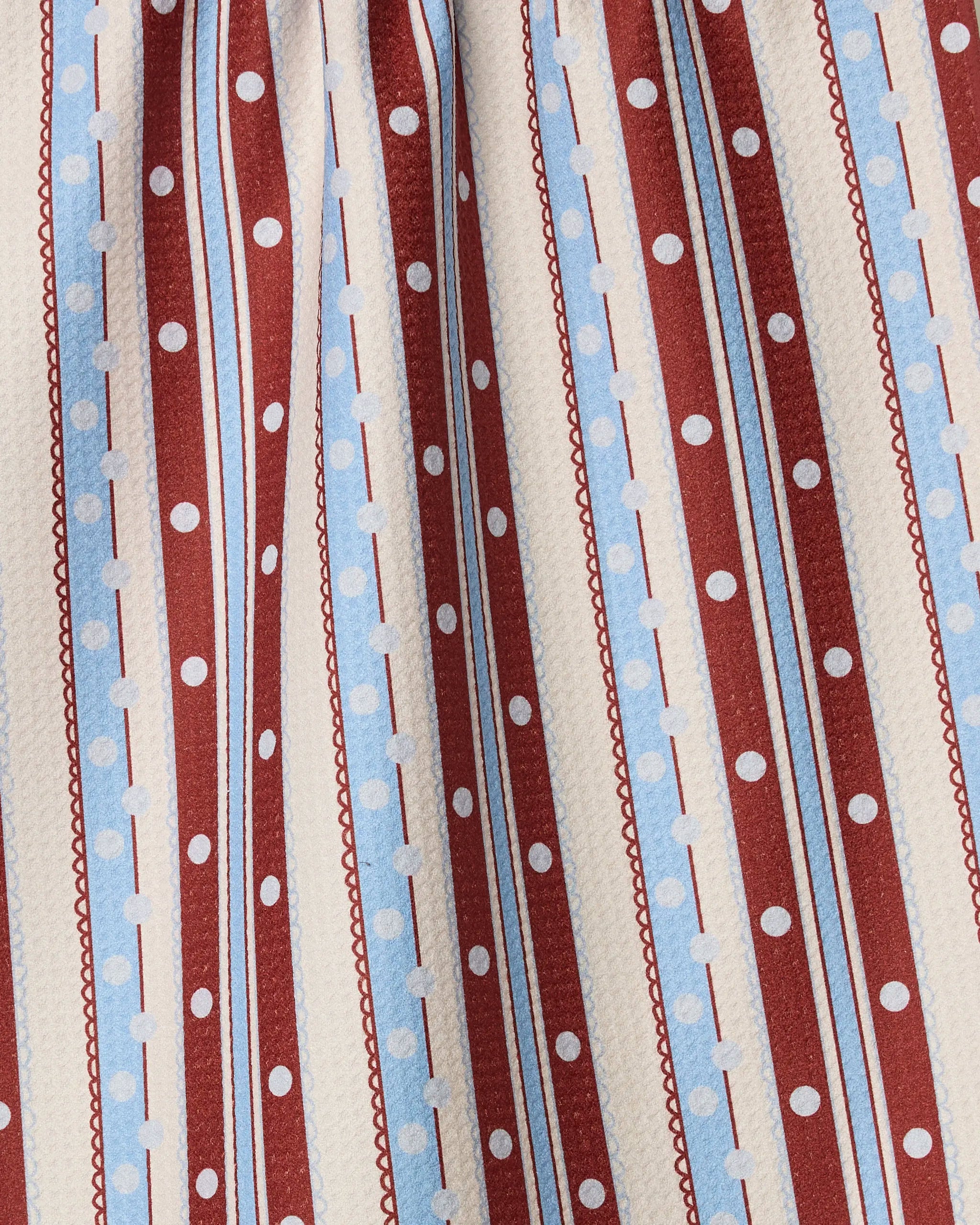 Close-up of Geometry's Blue Skies Stripes kitchen towel, made from recycled materials, with vertical red and white stripes, light blue dotted lines, white polka dots on the red stripes, and a slightly pleated fabric.