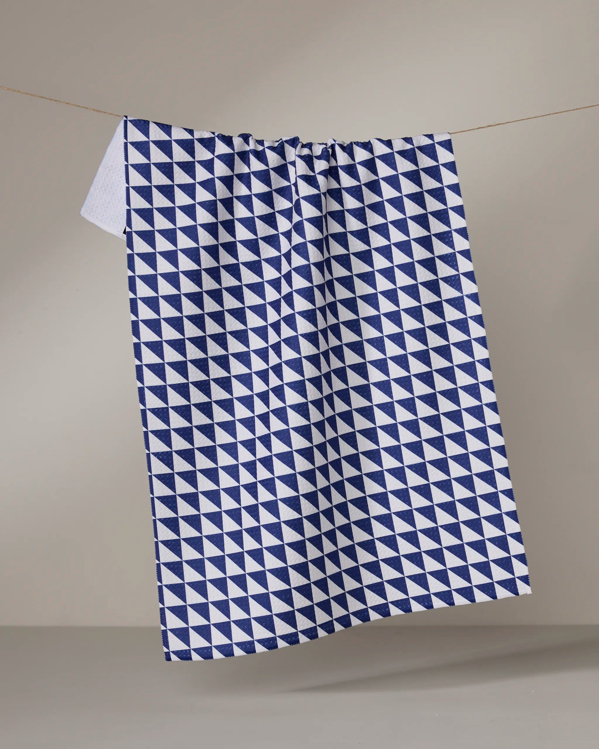 The Geometry Blue Whirl tea towel, featuring a blue and white geometric triangle pattern, hangs on a string against a plain light gray background. It’s quick-drying and stylish for any kitchen.