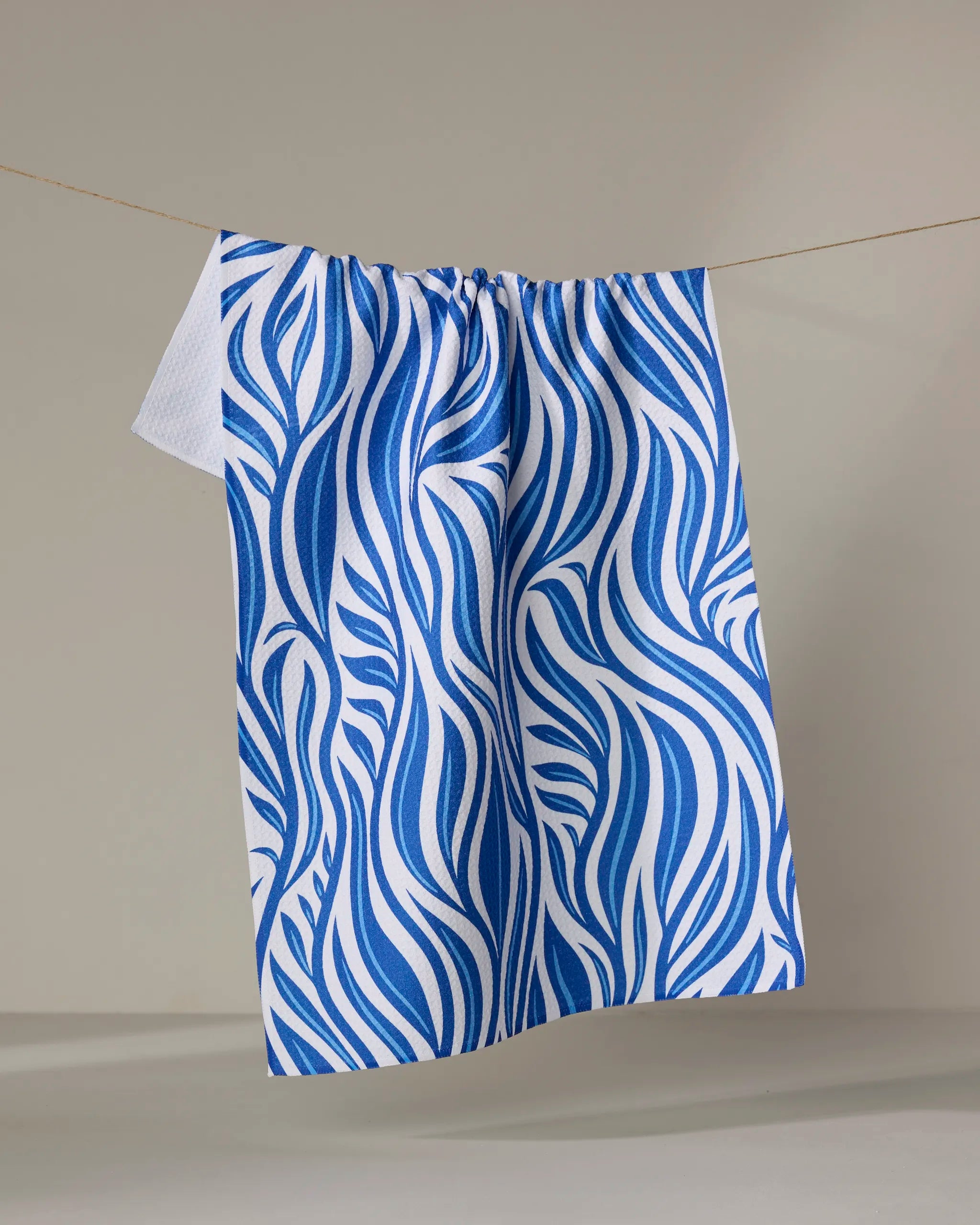 The Rachel Parker Blue Willow Wave recycled kitchen towel, featuring a bold blue wavy leaf pattern, hangs on a thin clothesline against a neutral background.