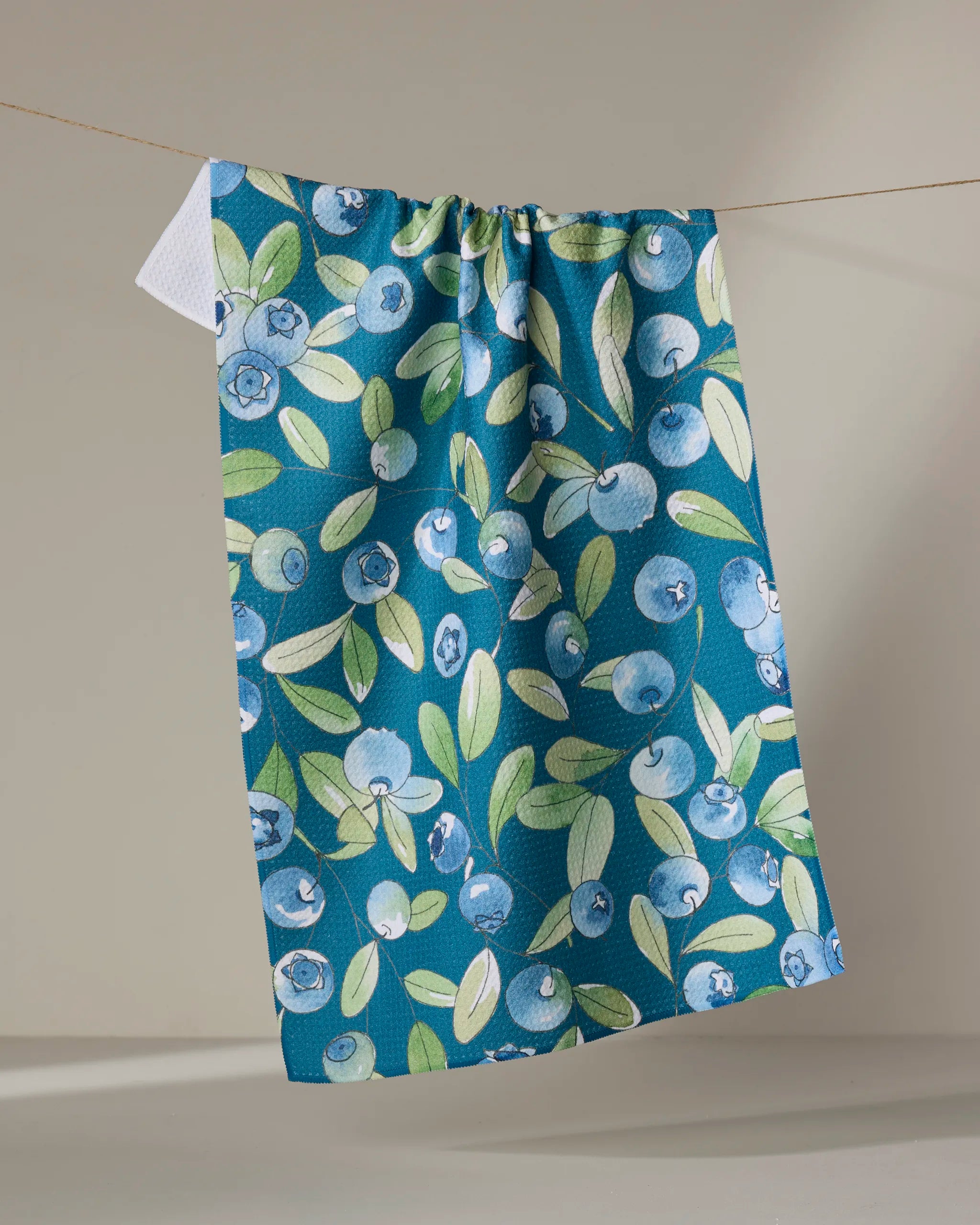 The "Blueberry Hill" tea towel by Sara Mari features a blueberry and green leaf design, made from recycled materials. It hangs on a clothesline against a neutral background.