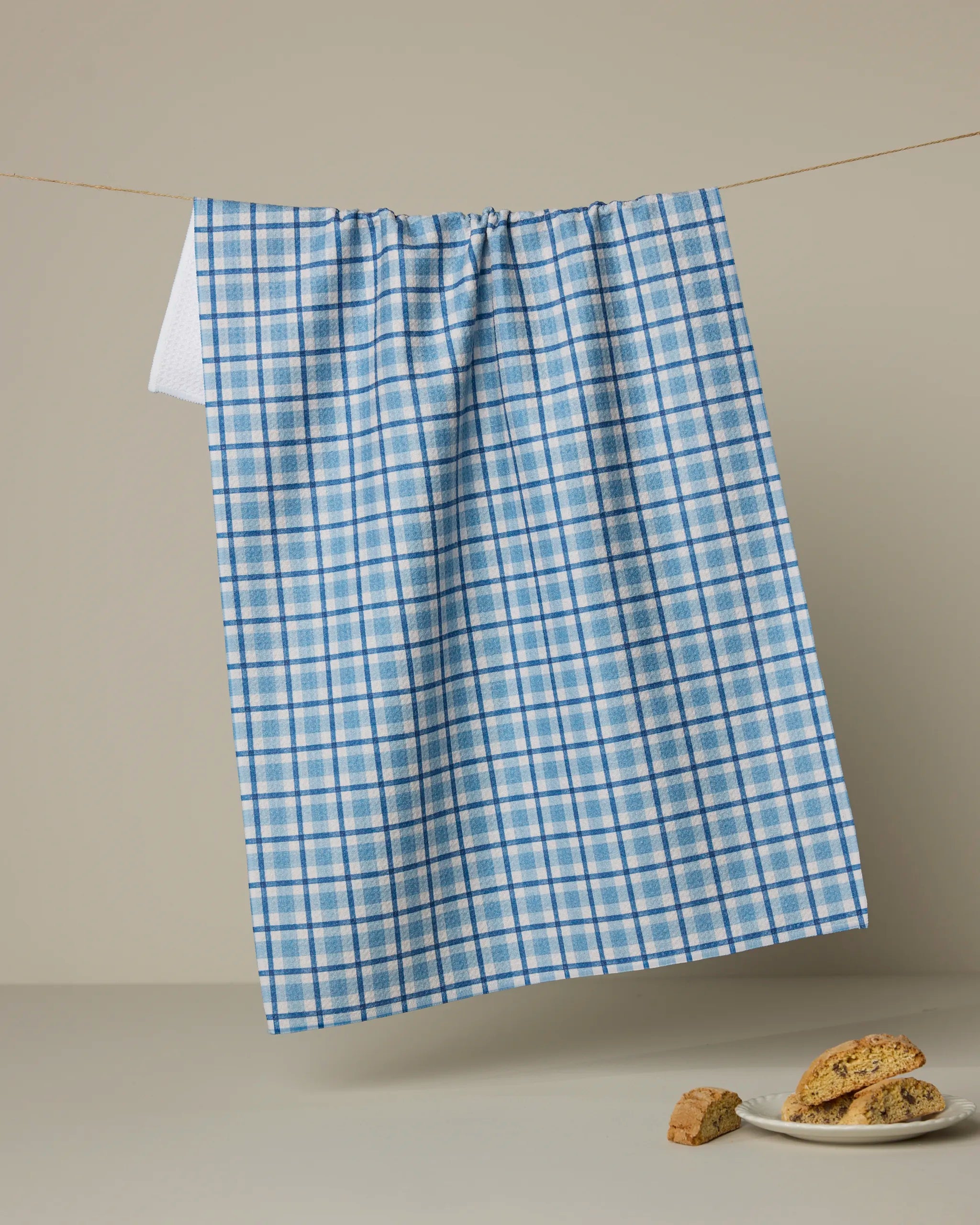 The Geometry Bluefield tea towel, featuring a blue and white check pattern, hangs on a line against a beige background with biscotti pieces and a white plate arranged below.
