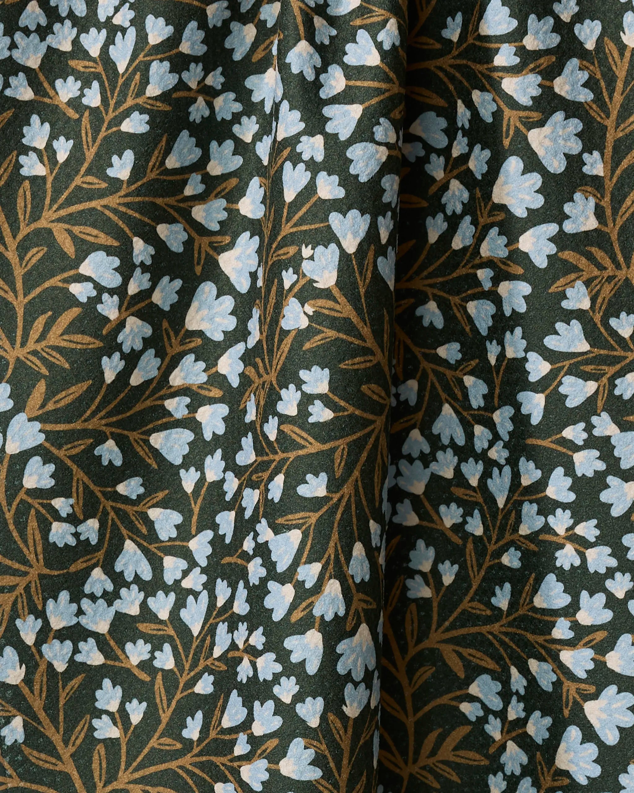 A close-up of Danelys Sidron’s Bluewood Garden tea towel shows small light blue flowers with brown stems on a dark green background. Made from recycled materials, it drapes softly, creating gentle folds and shadows.