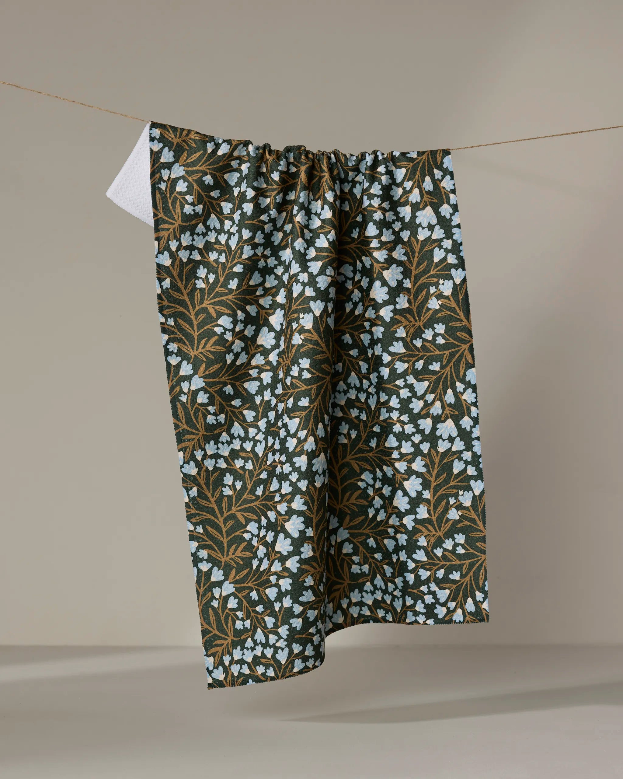 The "Bluewood Garden" tea towel by Danelys Sidron features light blue flowers and brown branches on a dark fabric, hanging on a clothesline against a plain, light background.