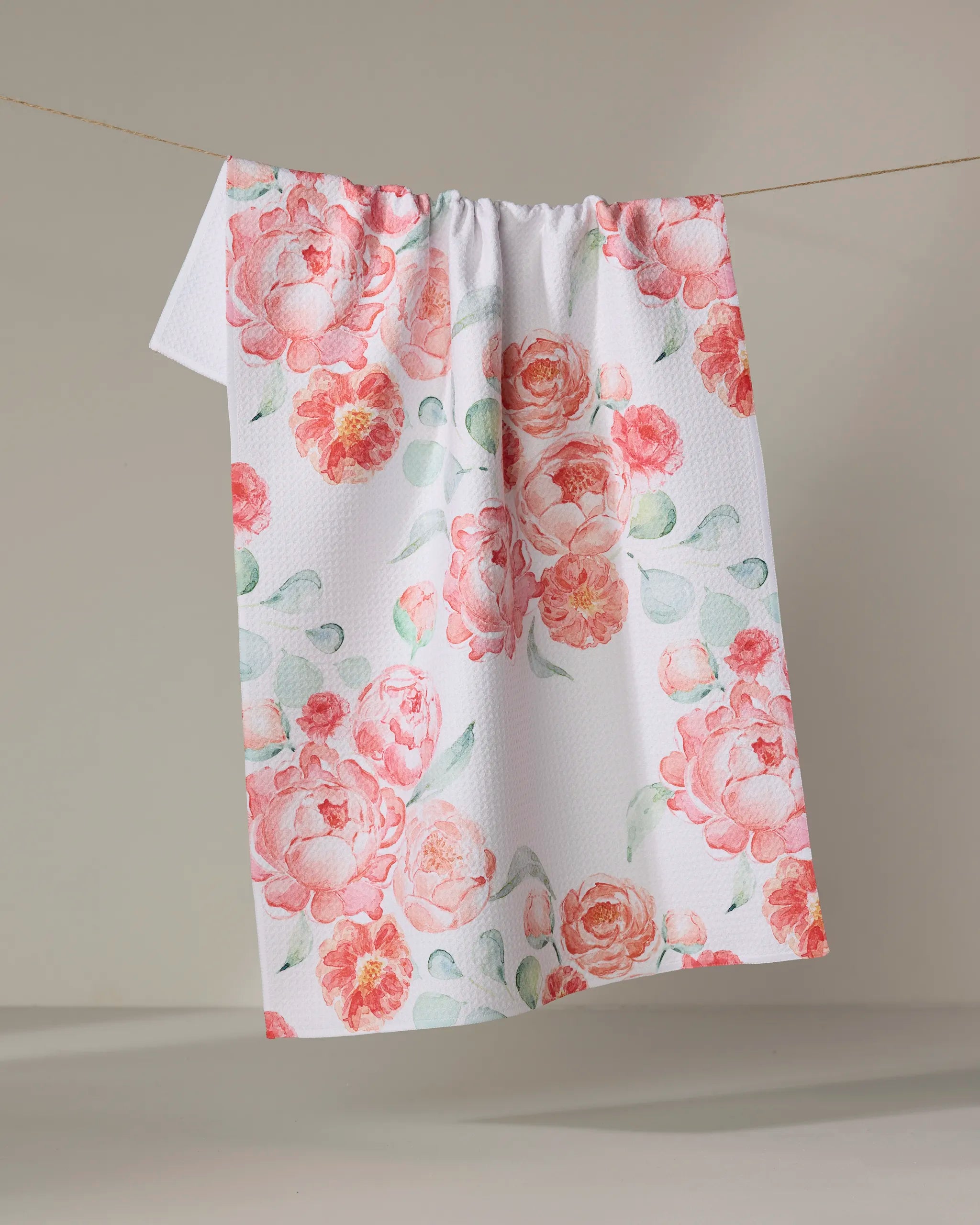 The "Blush Florals" kitchen towel by Elena Fay displays pink and peach watercolor-style flowers with green leaves on a white, textured fabric, hanging on a line against a neutral backdrop.