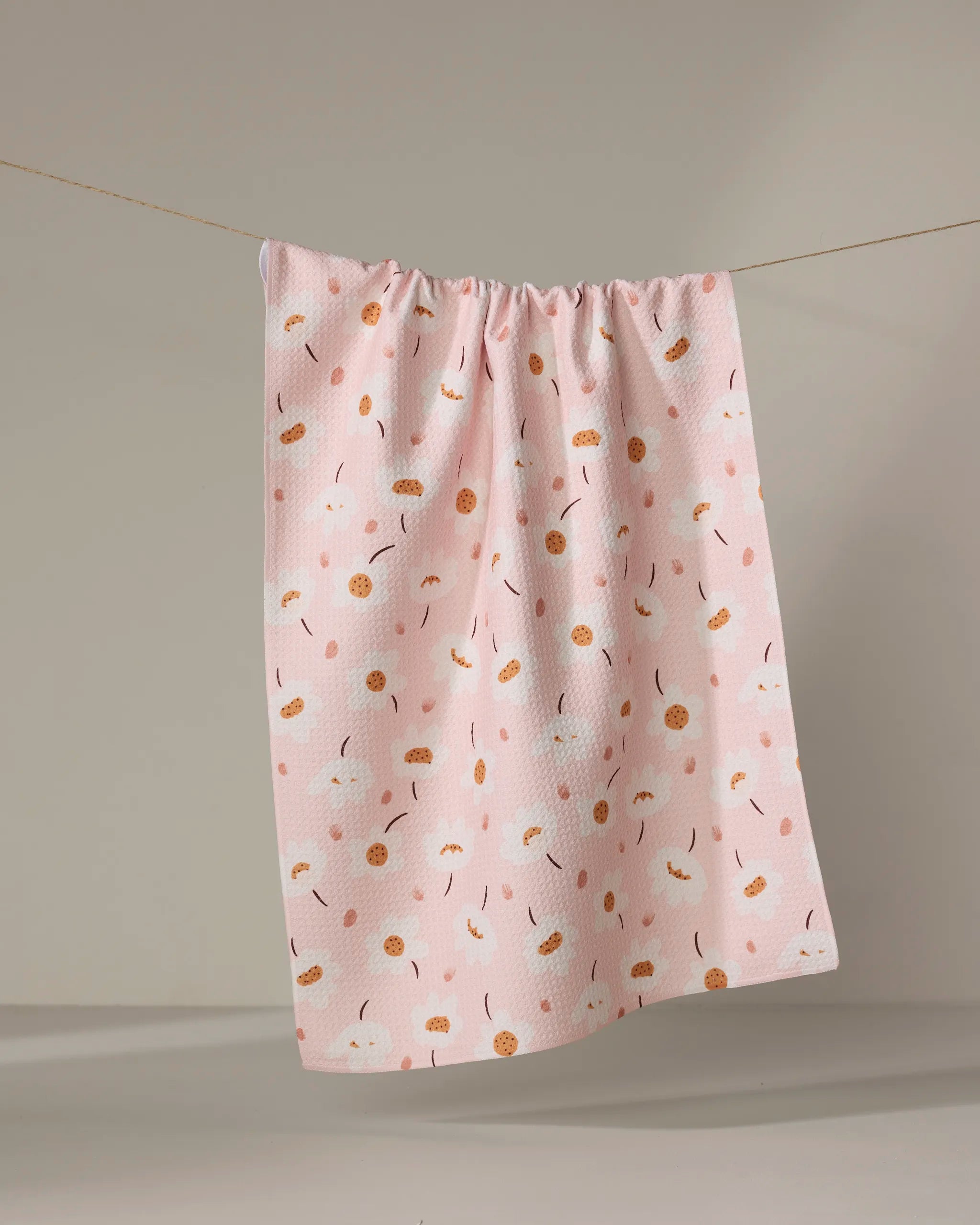 The Sara Mari "Blush Meadows" tea towel, made from recycled materials and adorned with white and orange flowers, hangs on a clothesline against a neutral background.