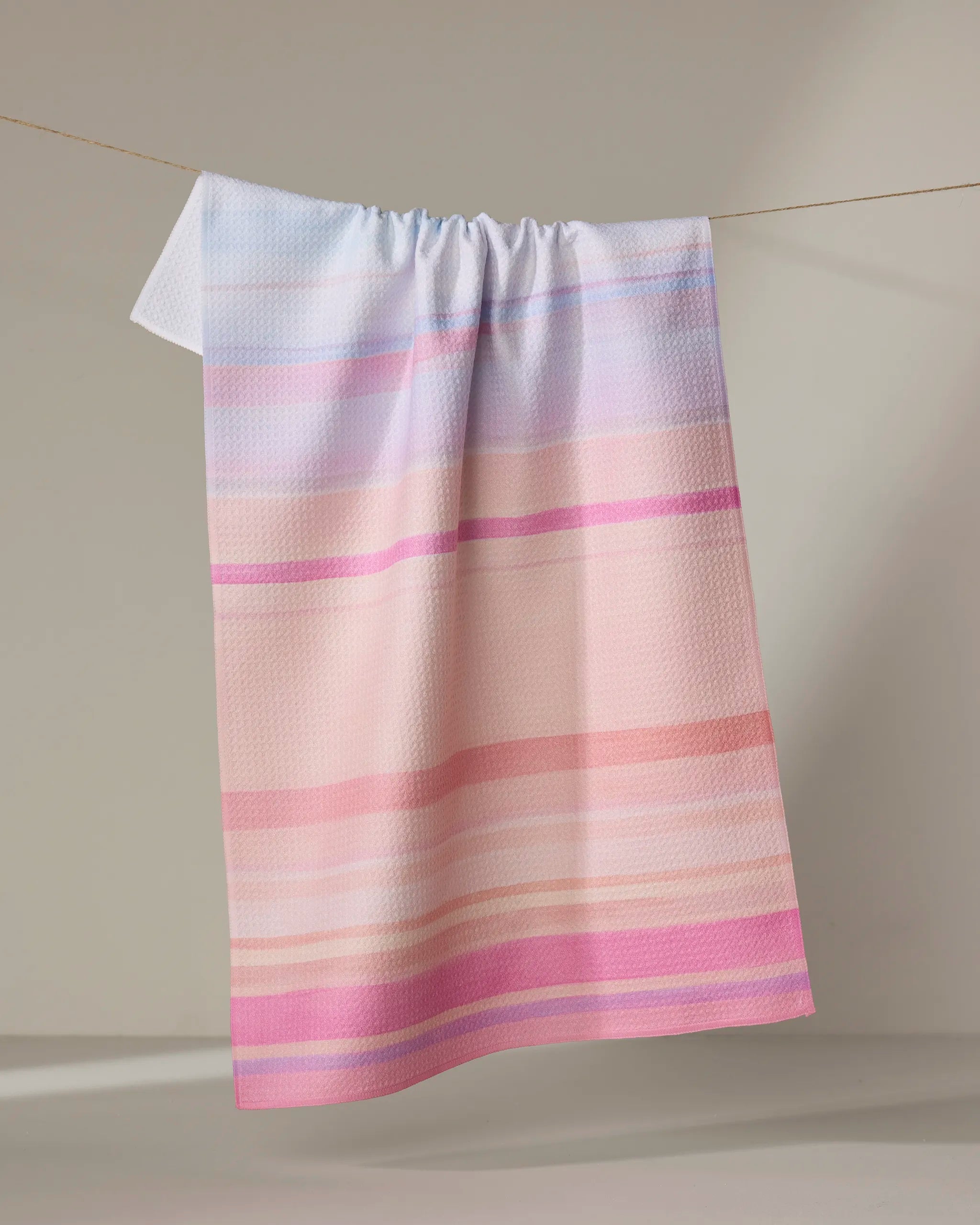 The Stefanie Bales "Blush" towel, featuring ultra-absorbent fabric and pastel horizontal stripes in pink, peach, and lavender, hangs on a clothesline against a neutral background.