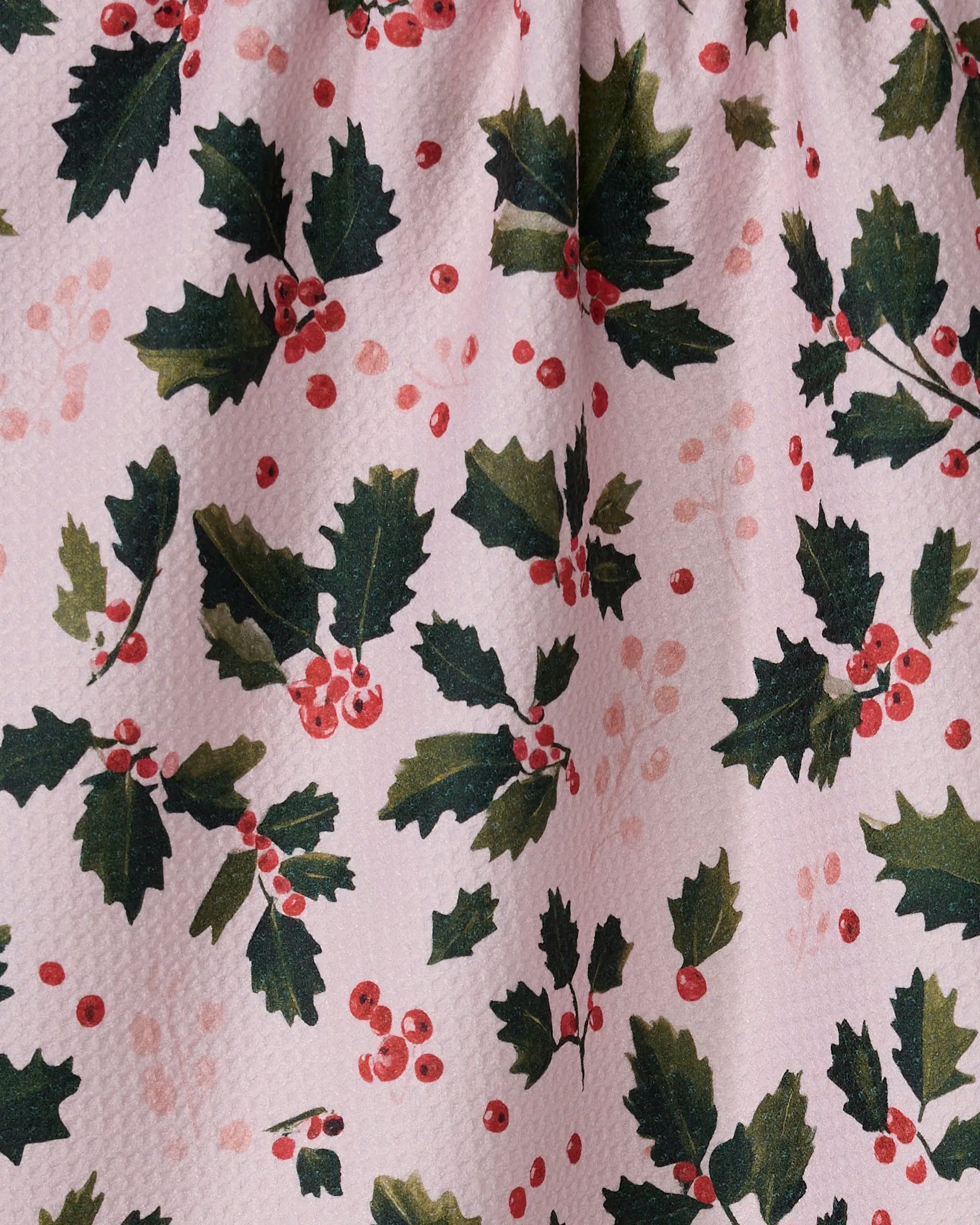 A close-up of Geometry's Blushberry tea towel shows green holly leaves and red berries on a light pink background. Made from recycled materials, its textured fabric and festive look bring holiday cheer to any kitchen.