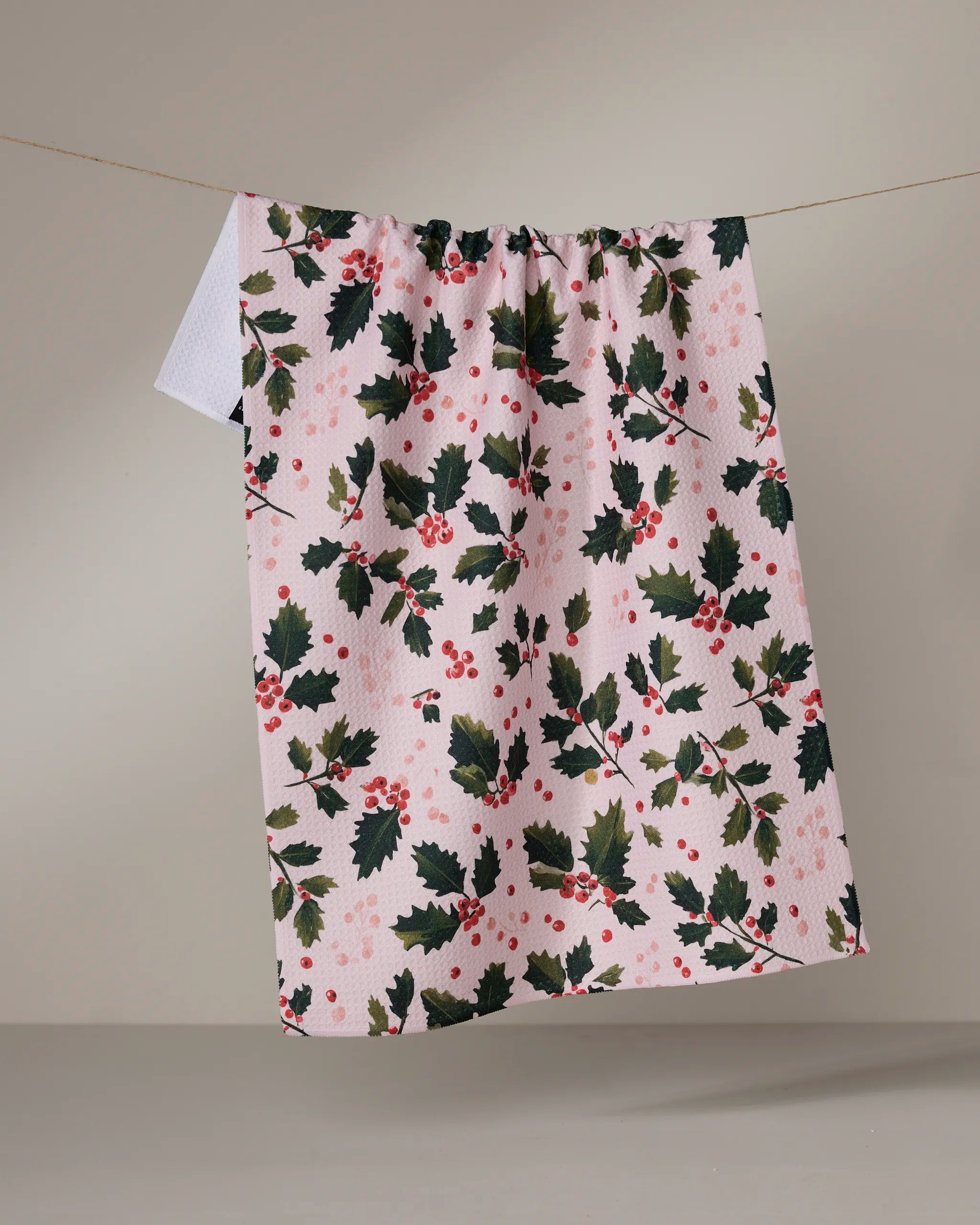 The Geometry Blushberry tea towel, made from recycled materials, hangs on a line with green holly and red berries on a pink background—festive, ultra-absorbent, and eco-friendly.