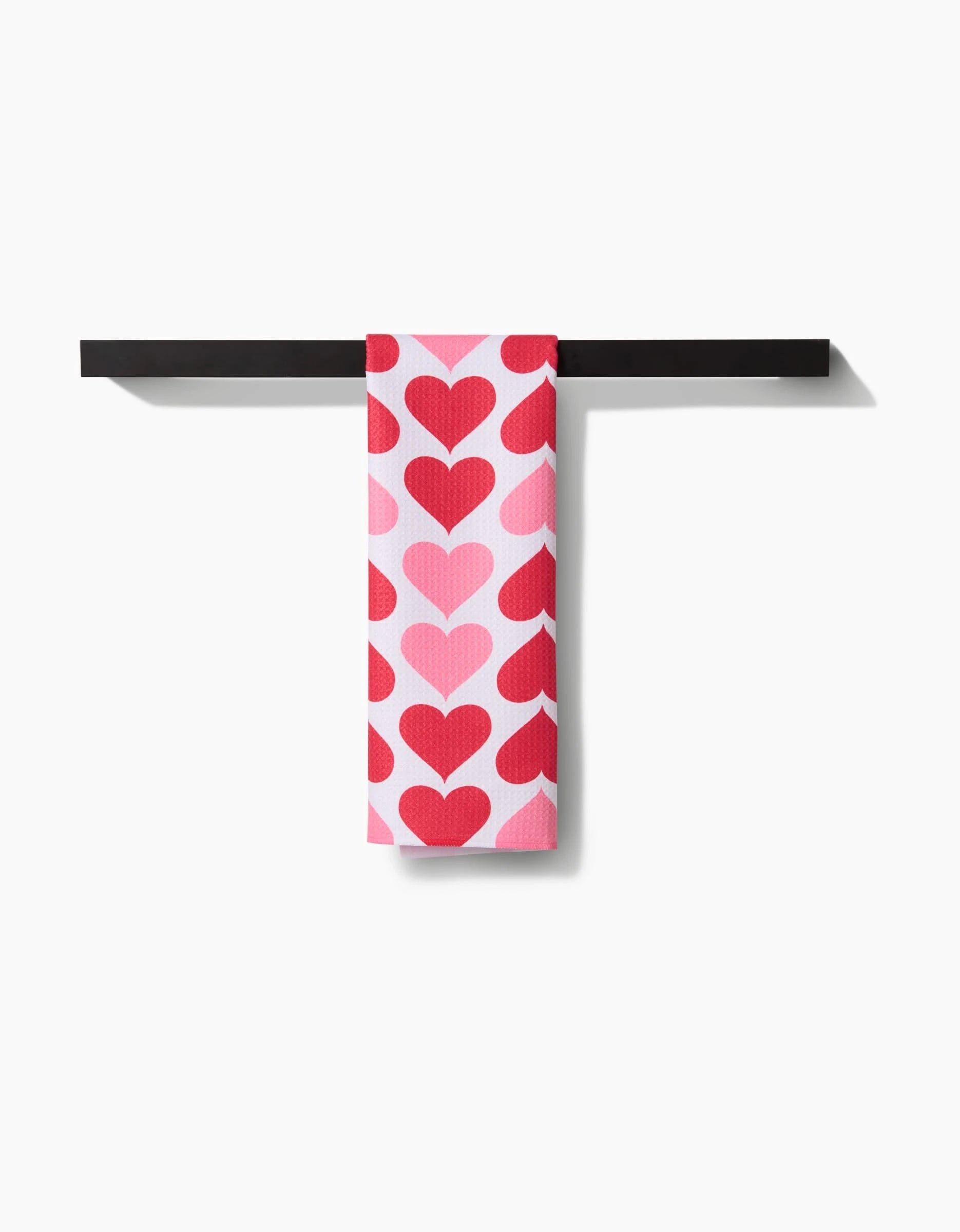 The Danelys Sidron Blushing Hearts towel, featuring red and pink heart patterns, hangs neatly on a black towel bar against a white background.