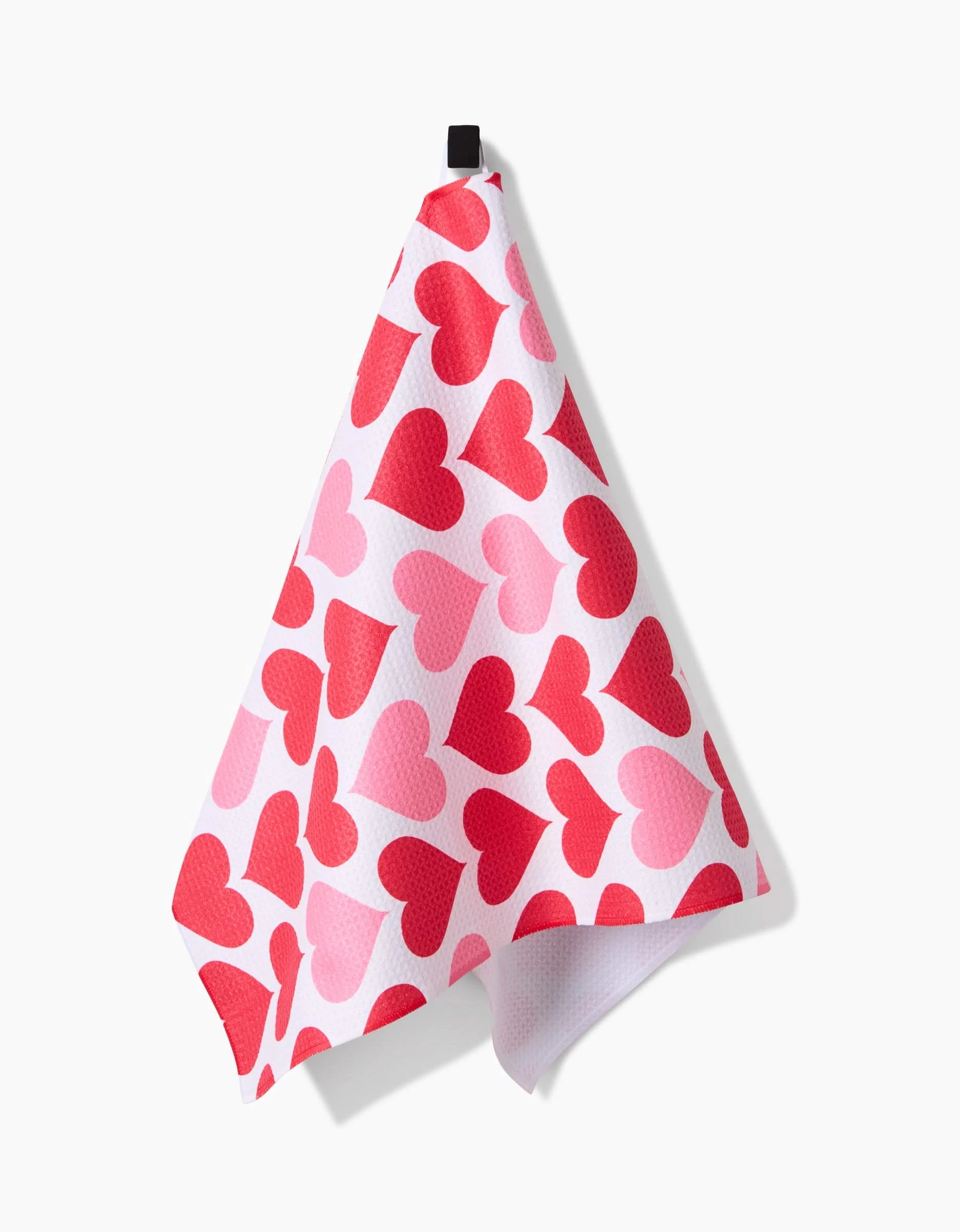 The Danelys Sidron Blushing Hearts tea towel, featuring pink and red heart patterns, hangs from a black hook against a plain white background.