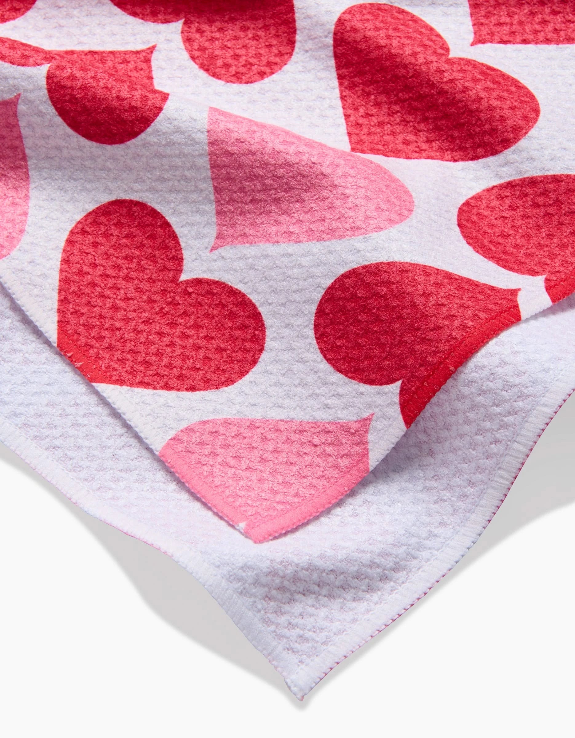 A close-up of Danelys Sidrons Blushing Hearts tea towel, ultra-absorbent and white, adorned with large red and pink heart patterns; one corner is folded to display the underside.