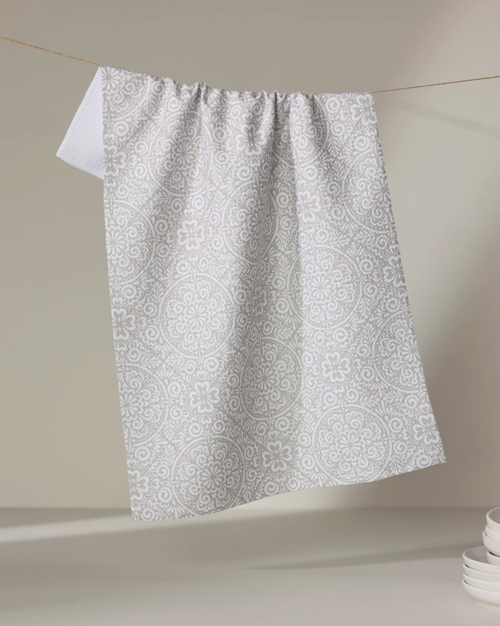 The Kris Ruff Boho Medallion dish towel, featuring a white and light grey pattern, hangs on a string against a neutral background, with a stack of white plates in the lower right corner.