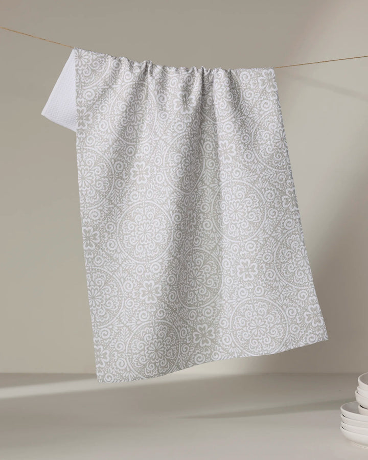 The Kris Ruff Boho Medallion dish towel, featuring a white and light grey pattern, hangs on a string against a neutral background, with a stack of white plates in the lower right corner.