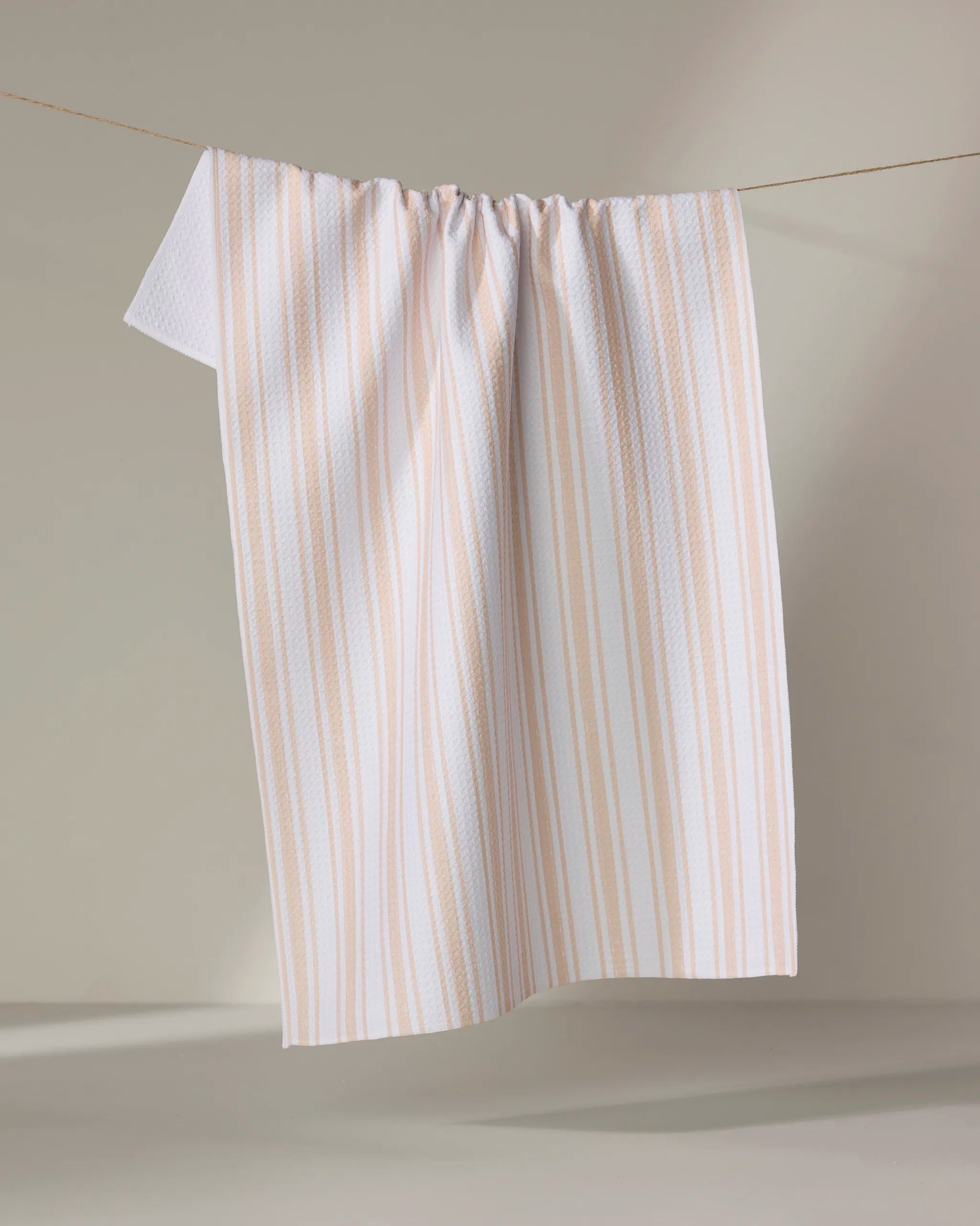 The Boho Stripe towel by Hufton Studio, featuring white fabric with subtle beige vertical stripes, hangs over a thin clothesline against a plain, light background.