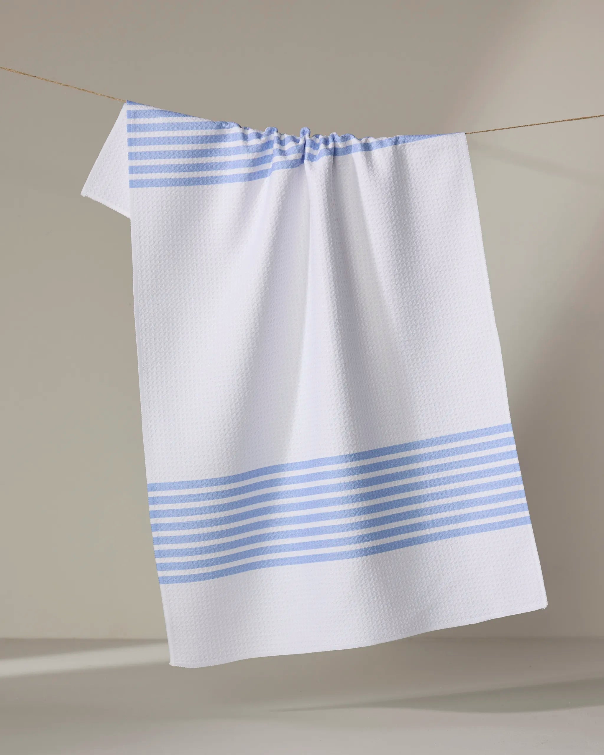 The Buy Guide’s Bold Blank Space - French Blue tea towel, made from recycled materials, features a textured white fabric with two sets of horizontal light blue stripes and hangs on a line against a neutral background.