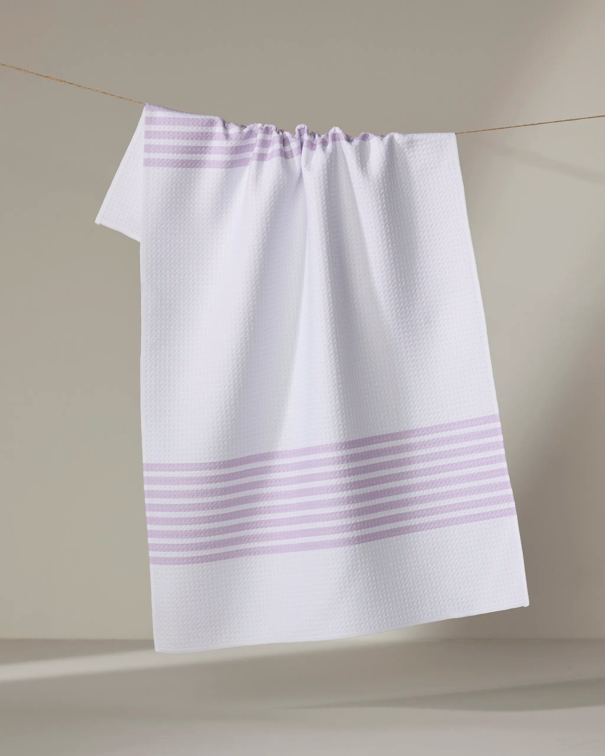 The Buy Guide’s Bold Blank Space - Lilac tea towel, featuring light purple stripes on ultra-absorbent white fabric, hangs neatly on a clothesline against a neutral background.