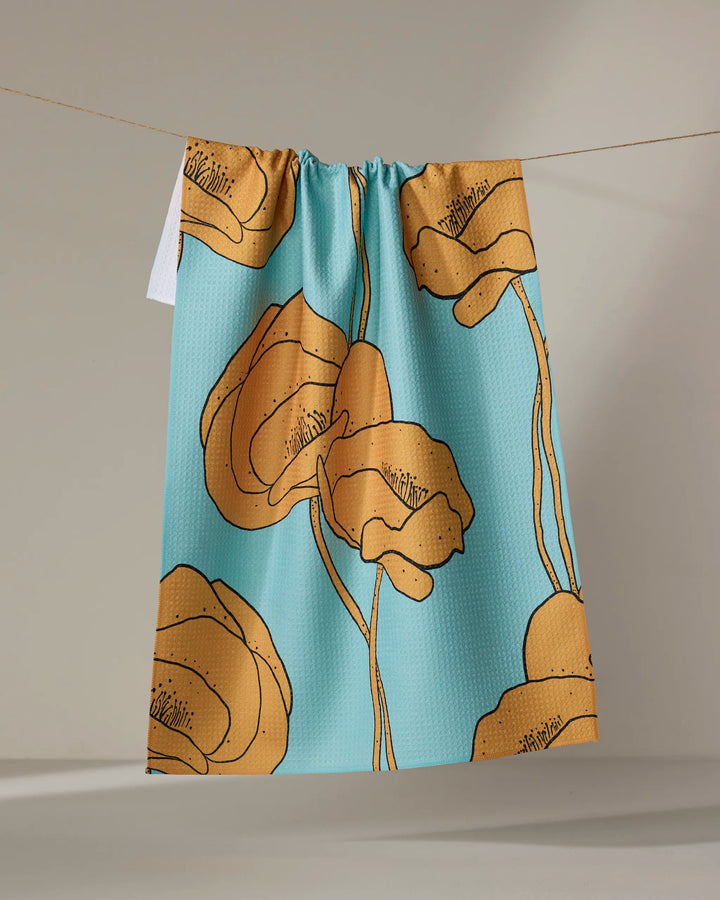 The Julianne Haness "Bold Peonies" tea towel, featuring a quick-drying fabric with a blue background and large stylized orange peony flowers, hangs on a line against a neutral backdrop.