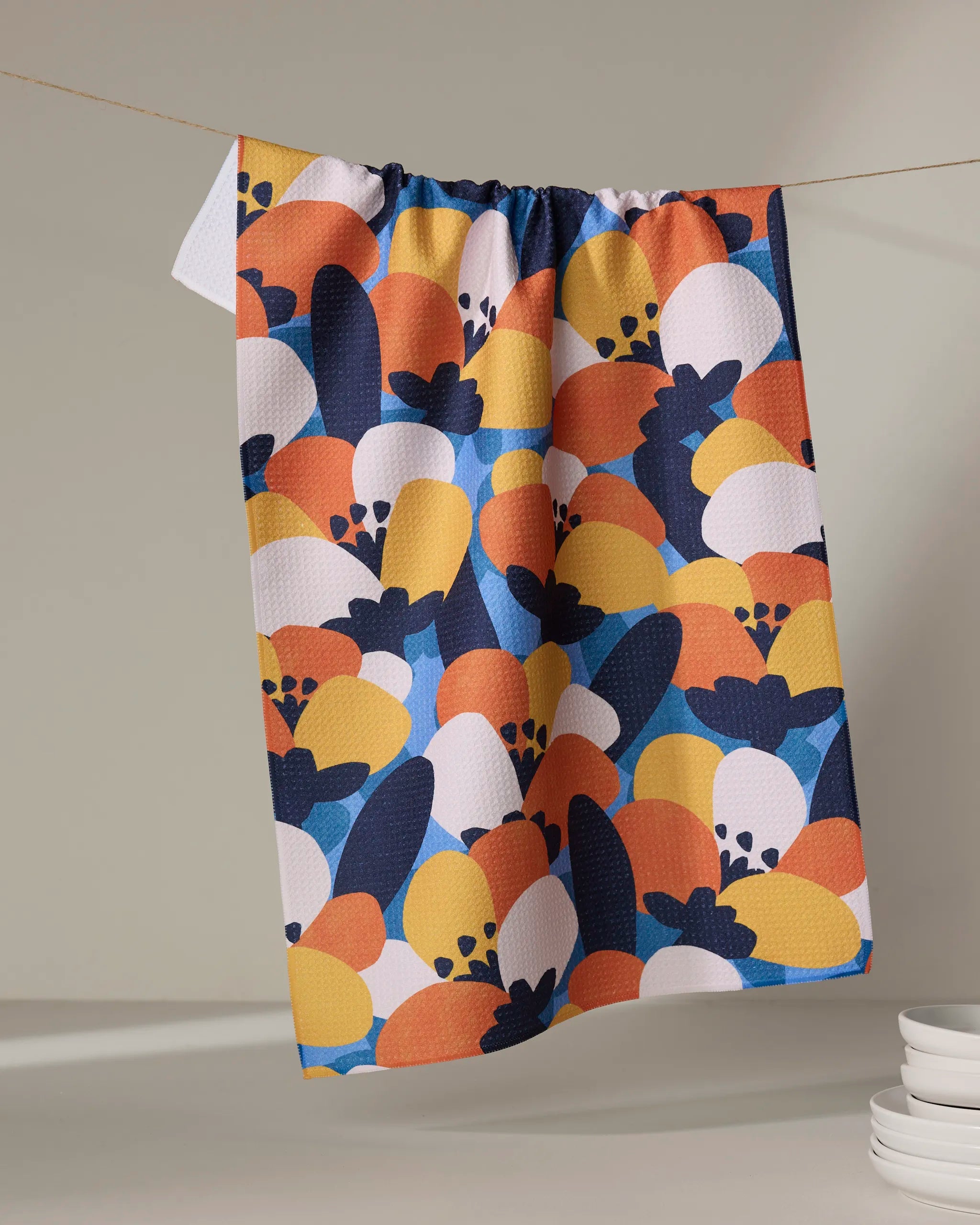 The Bold Petal Bloom tea towel by Claire Iglesias, made from recycled materials, features an abstract floral design in blue, orange, yellow, navy, and white and hangs on a clothesline beside stacked white dishes on a light surface.