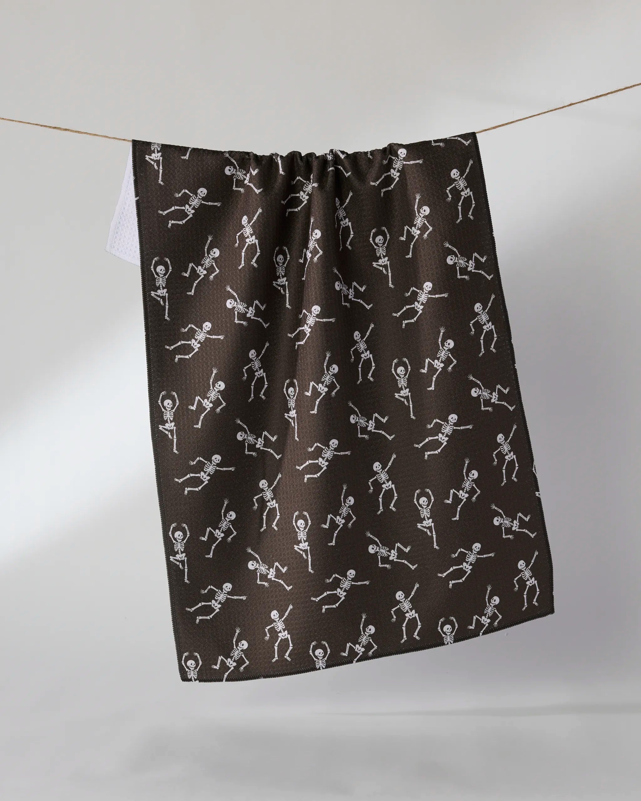 The Bone Jamboree by Geometry is a dark, ultra-absorbent towel made from recycled materials with a fun white stick figure pattern, shown hanging on a clothesline against a plain background.