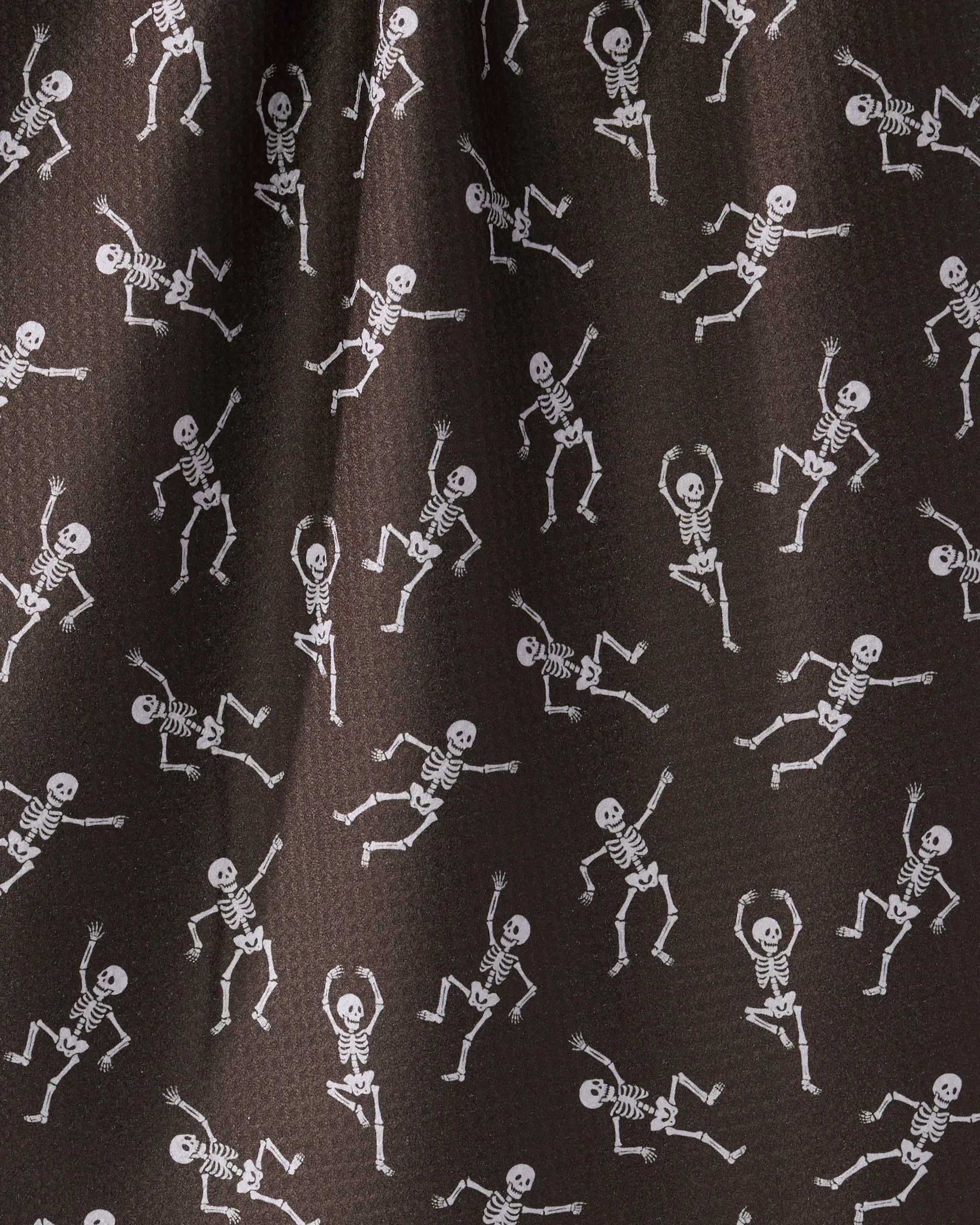 The Bone Jamboree tea towel by Geometry features playful white skeletons dancing in a repeating pattern on dark brown, slightly textured fabric made from recycled materials.