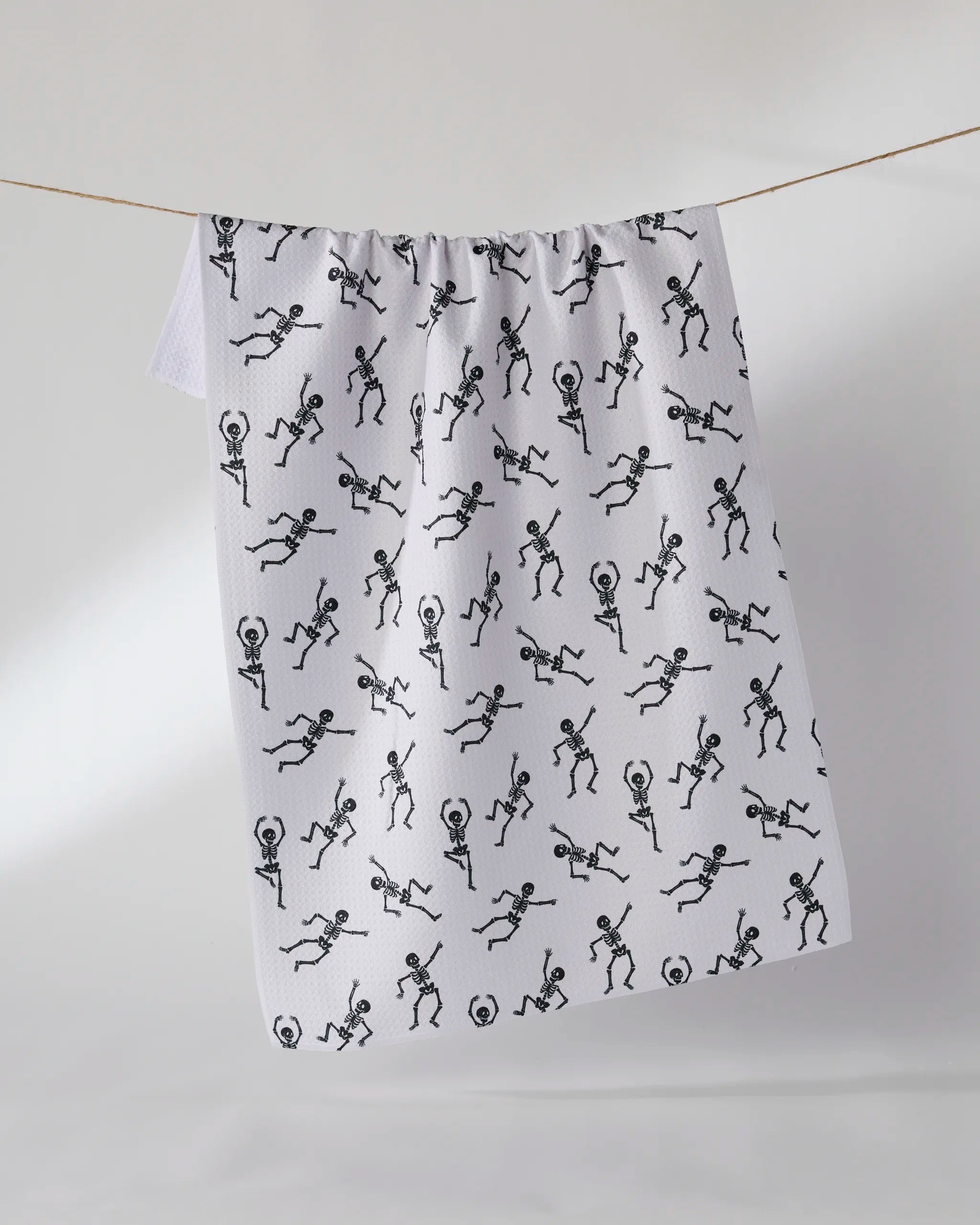 The Geometry Bone Jamboree tea towel, made from recycled materials, features black dancing skeletons on white and hangs on a clothesline against a light background.