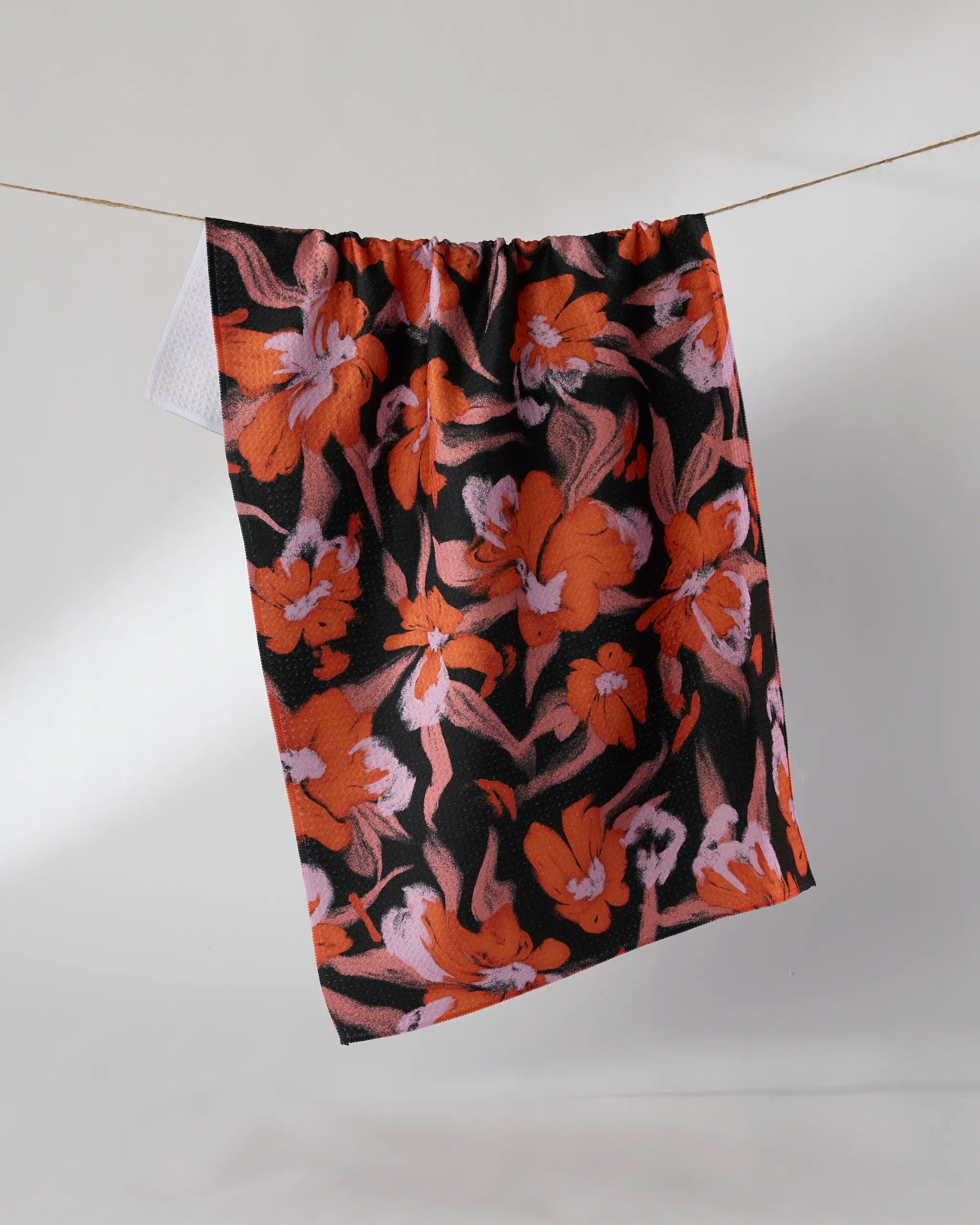 The Geometry Boo-tanical tea towel, featuring a vibrant orange and pink floral pattern on black, hangs on a string against a plain light gray background.