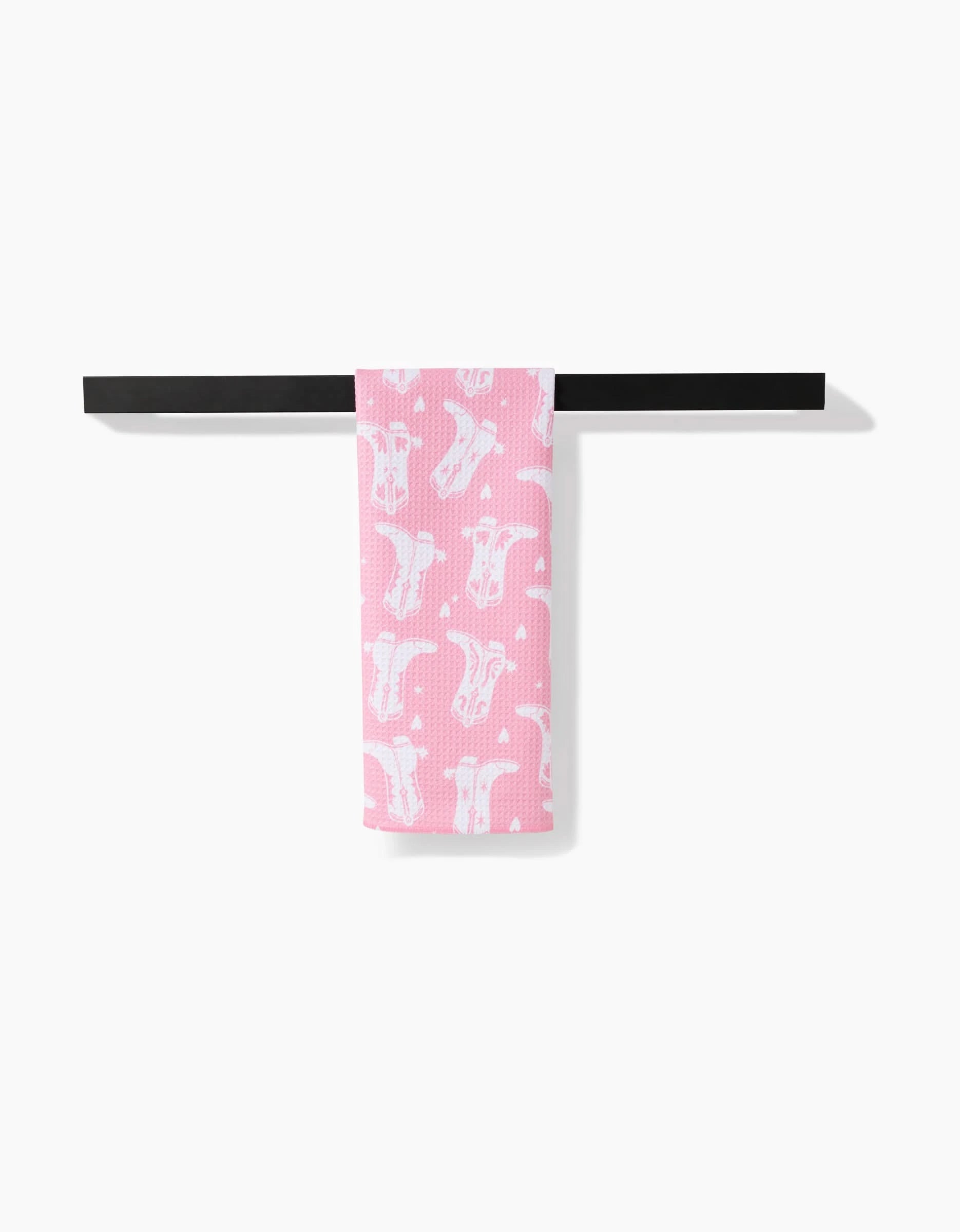 The Boots On Worries Gone tea towel by Aubrey Fairchild, featuring white cow face patterns on pink Geoweave fabric, hangs neatly over a black towel bar on a plain white background.
