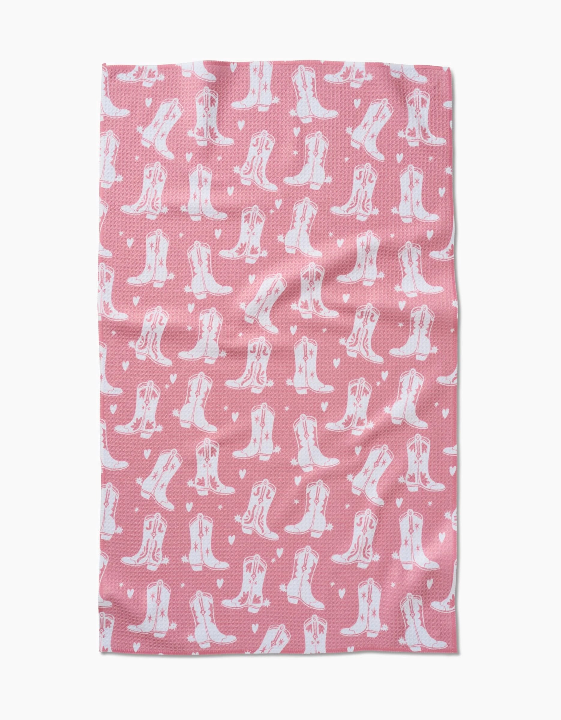 The “Boots On Worries Gone” tea towel by Aubrey Fairchild is made from Geoweave fabric and features a pink background with a repeating pattern of white cowboy boots and hearts.