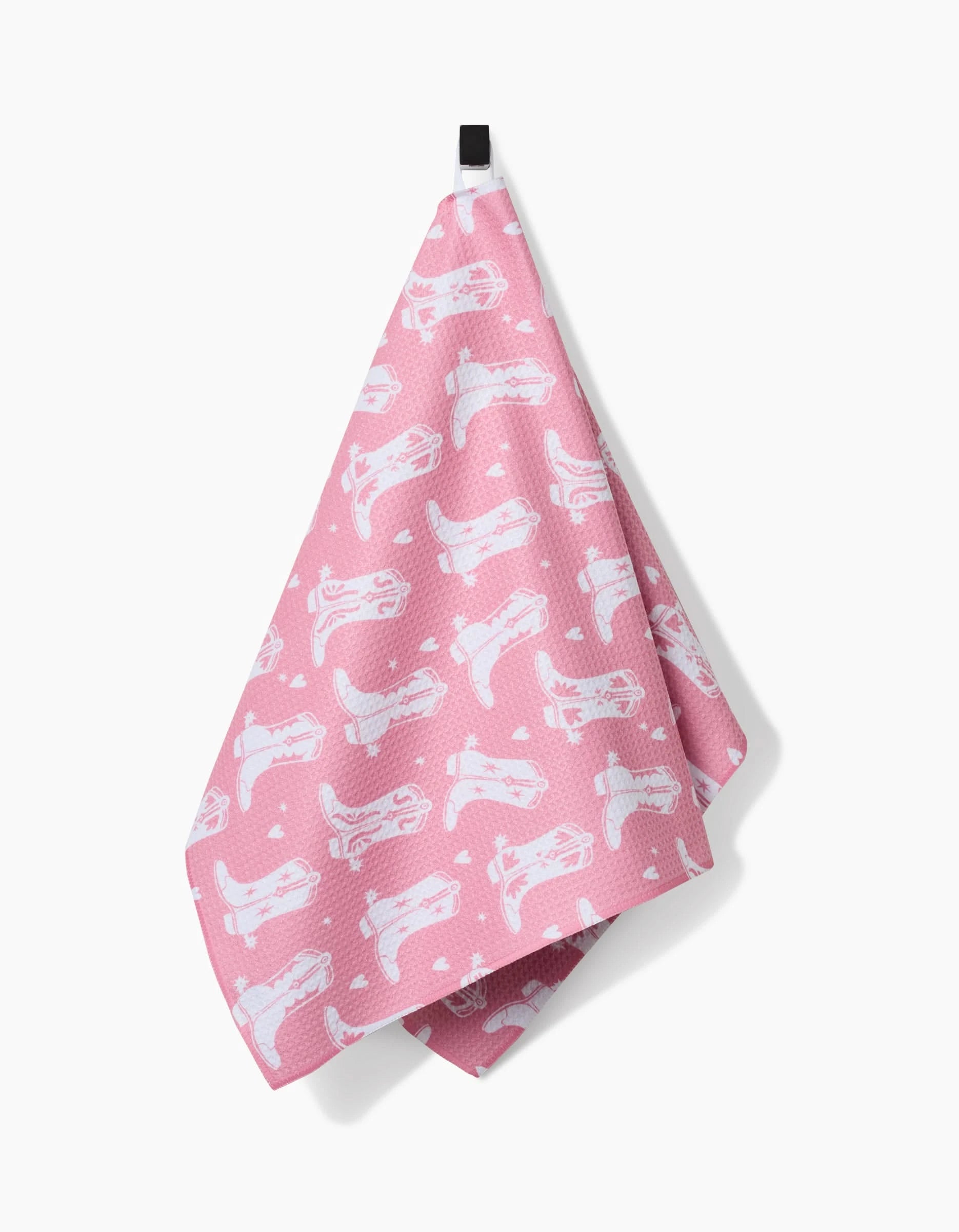 The “Boots On Worries Gone” Tea Towel by Aubrey Fairchild, featuring a repeating white cat pattern on pink and made from recycled materials, hangs on a black hook against a plain white background.