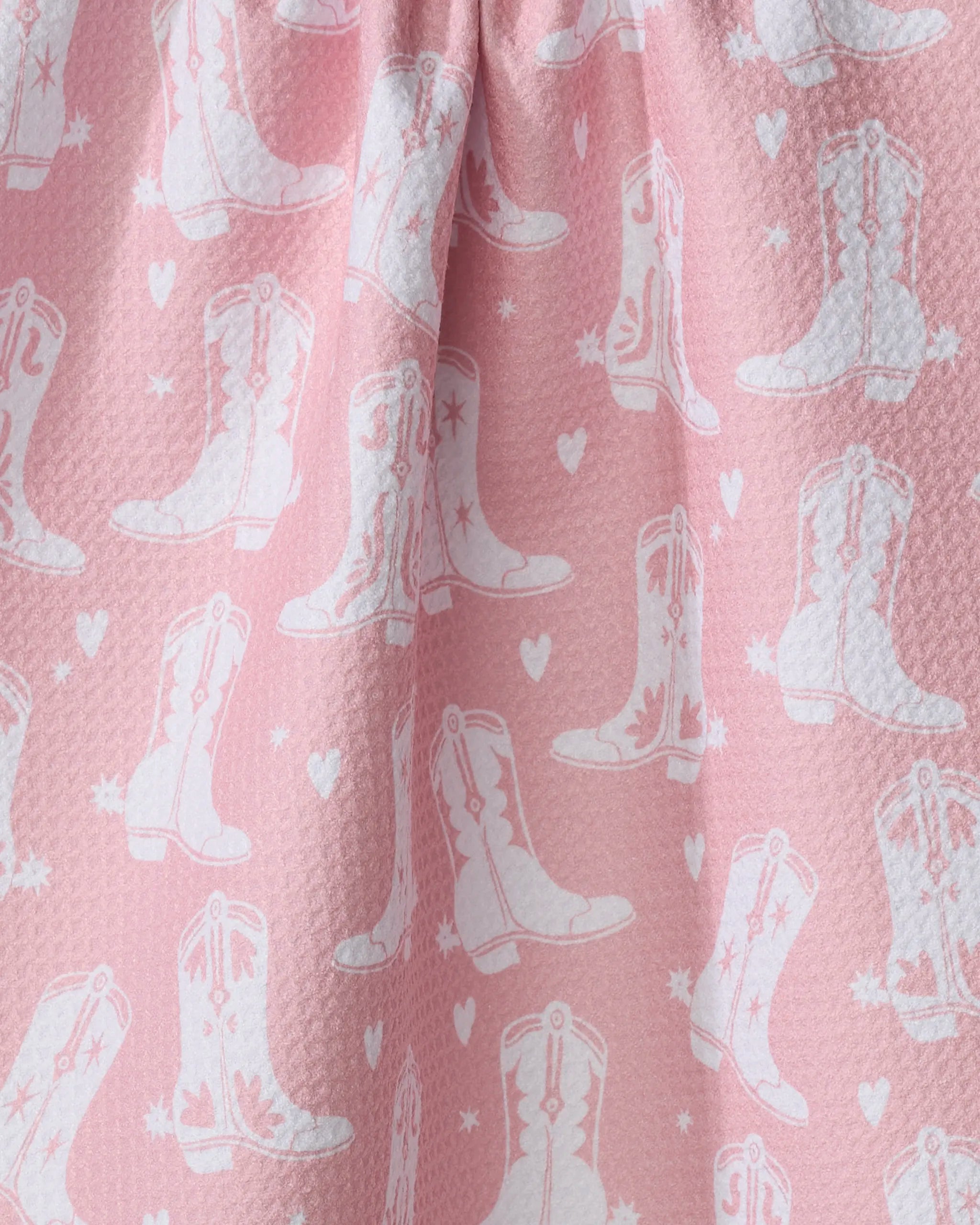 The "Boots On Worries Gone" kitchen towel by Aubrey Fairchild features pink textured fabric with white cowboy boots and hearts, softly gathered at the top—perfect for adding a playful, eco-friendly touch to your kitchen decor.