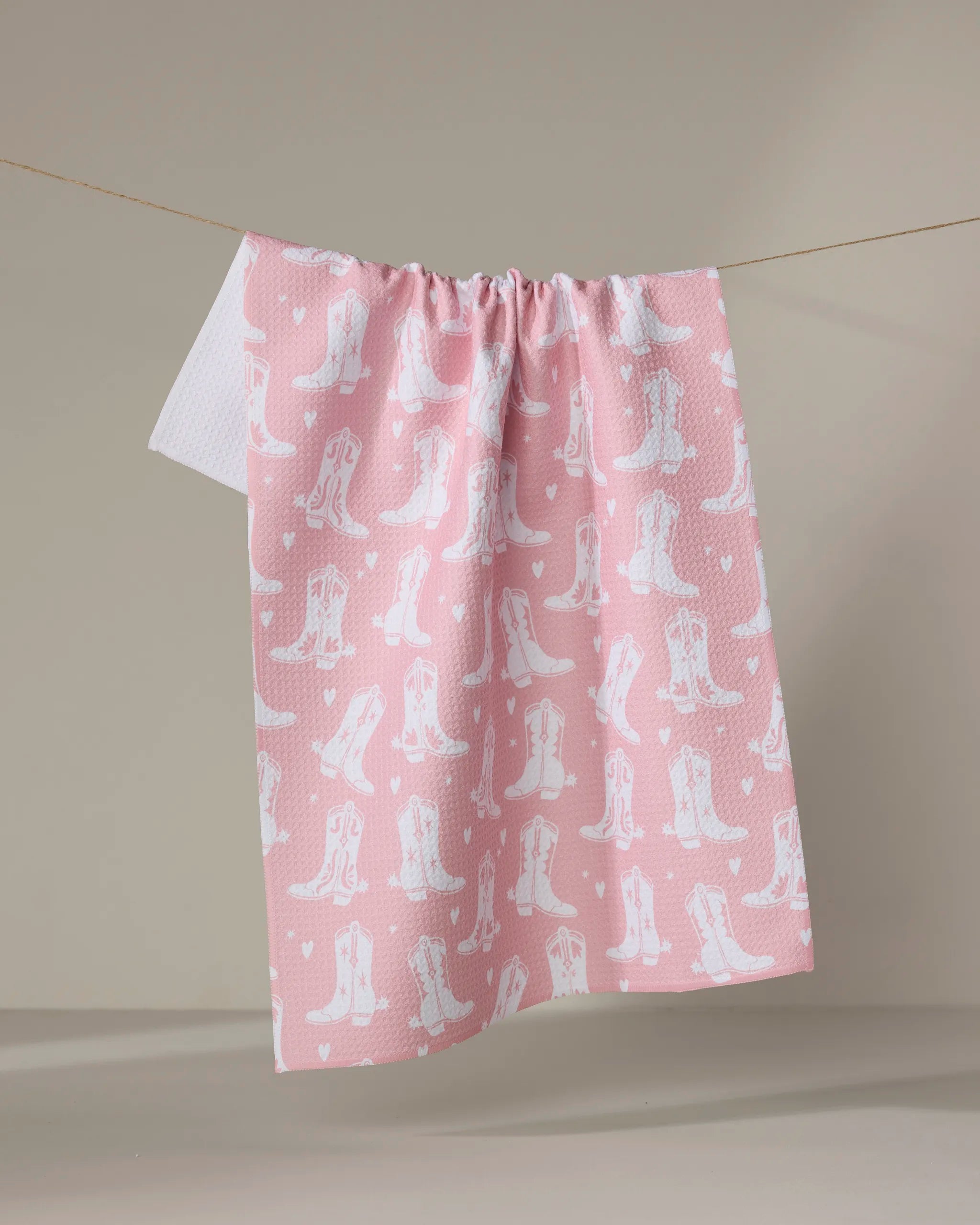 The "Boots On Worries Gone" tea towel by Aubrey Fairchild, featuring a pink eco-friendly fabric with white cowboy boots and hearts, hangs on a clothesline against a plain beige background.