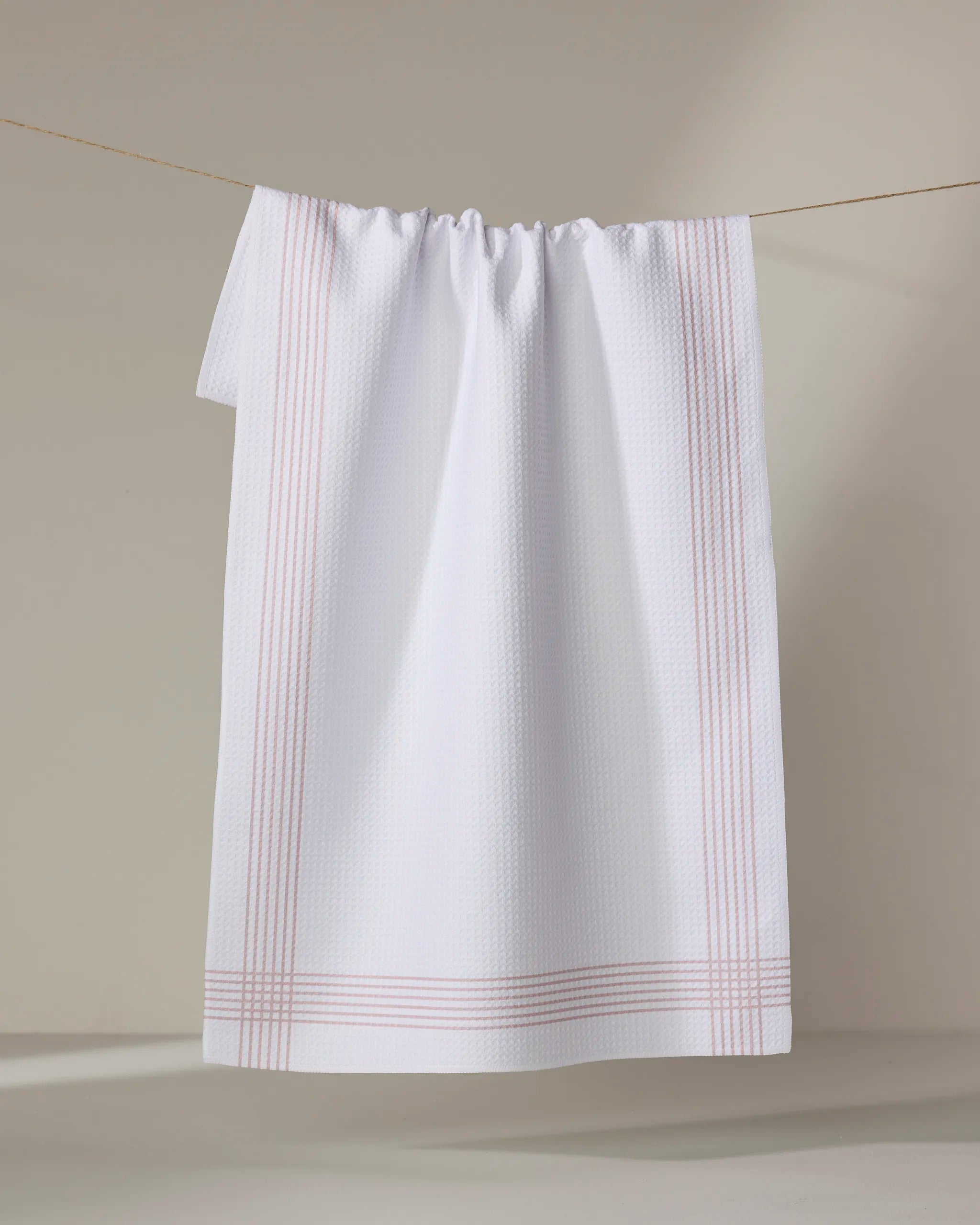 The Buy Guide's 'Borderline - Dusty Rose' is a white textured kitchen towel featuring light pink edge stripes, made from recycled materials, hanging on a clothesline against a neutral background.