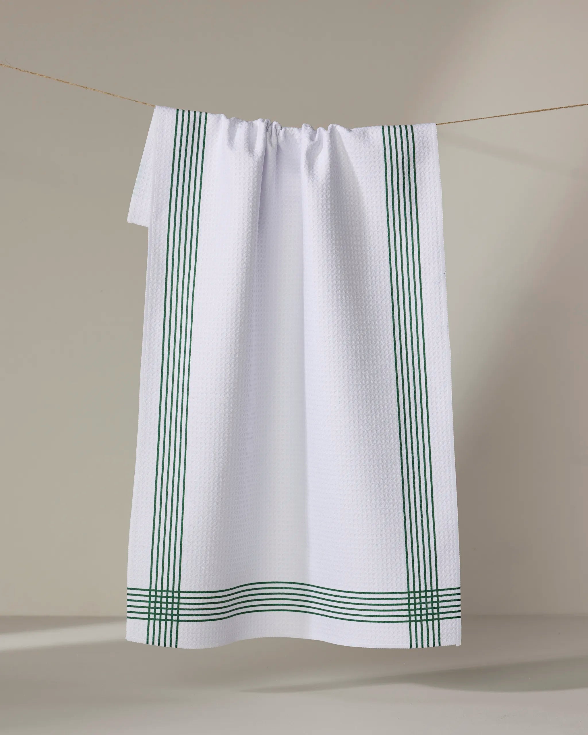 The Buy Guide’s Borderline - Evergreen tea towel, made from recycled materials, features a textured white design with green stripes and hangs on a string against a neutral background.