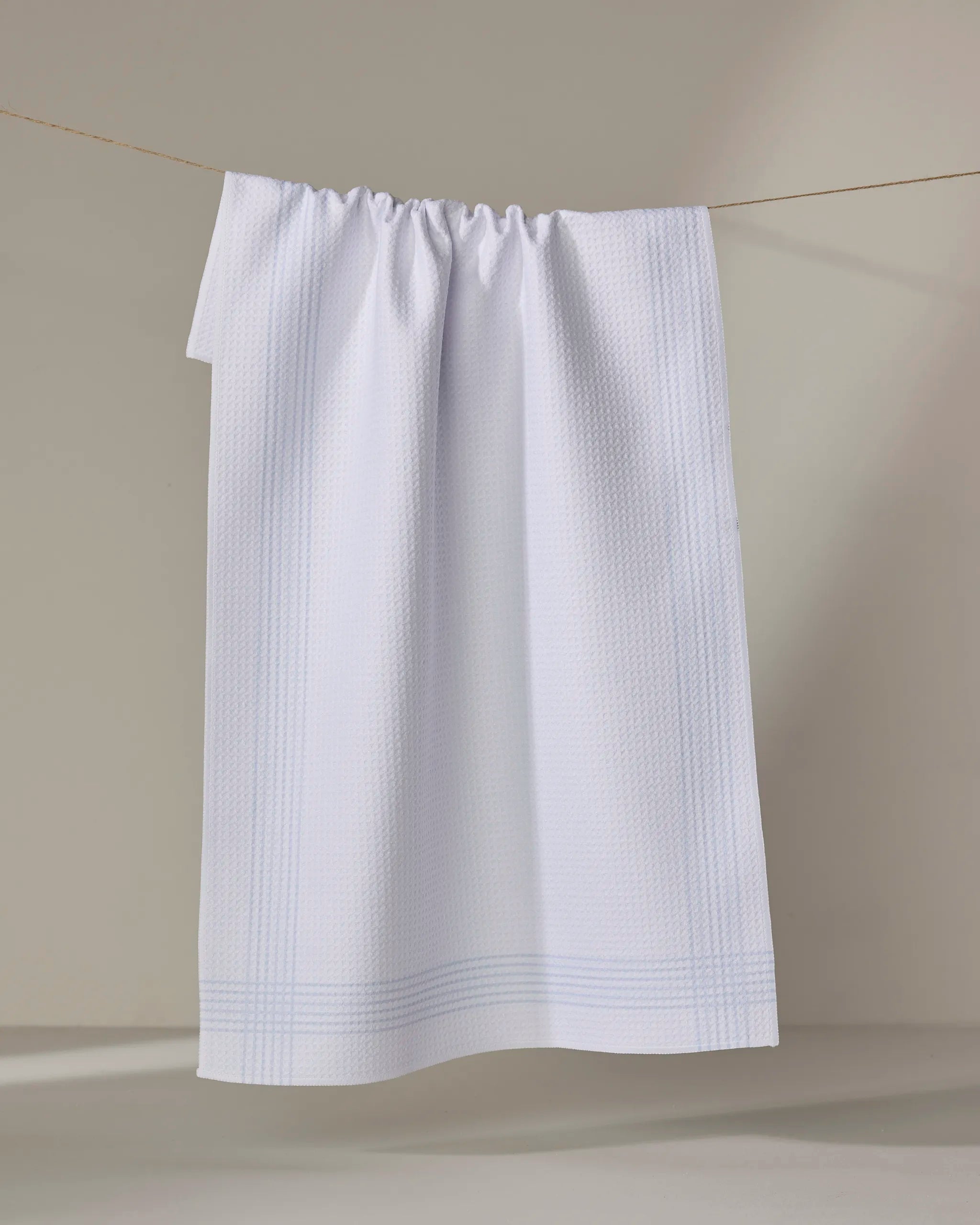 The Buy Guide’s Borderline - Light Blue tea towel, featuring a white textured design with subtle stripes and made from recycled materials, hangs on a clothesline against a neutral background.