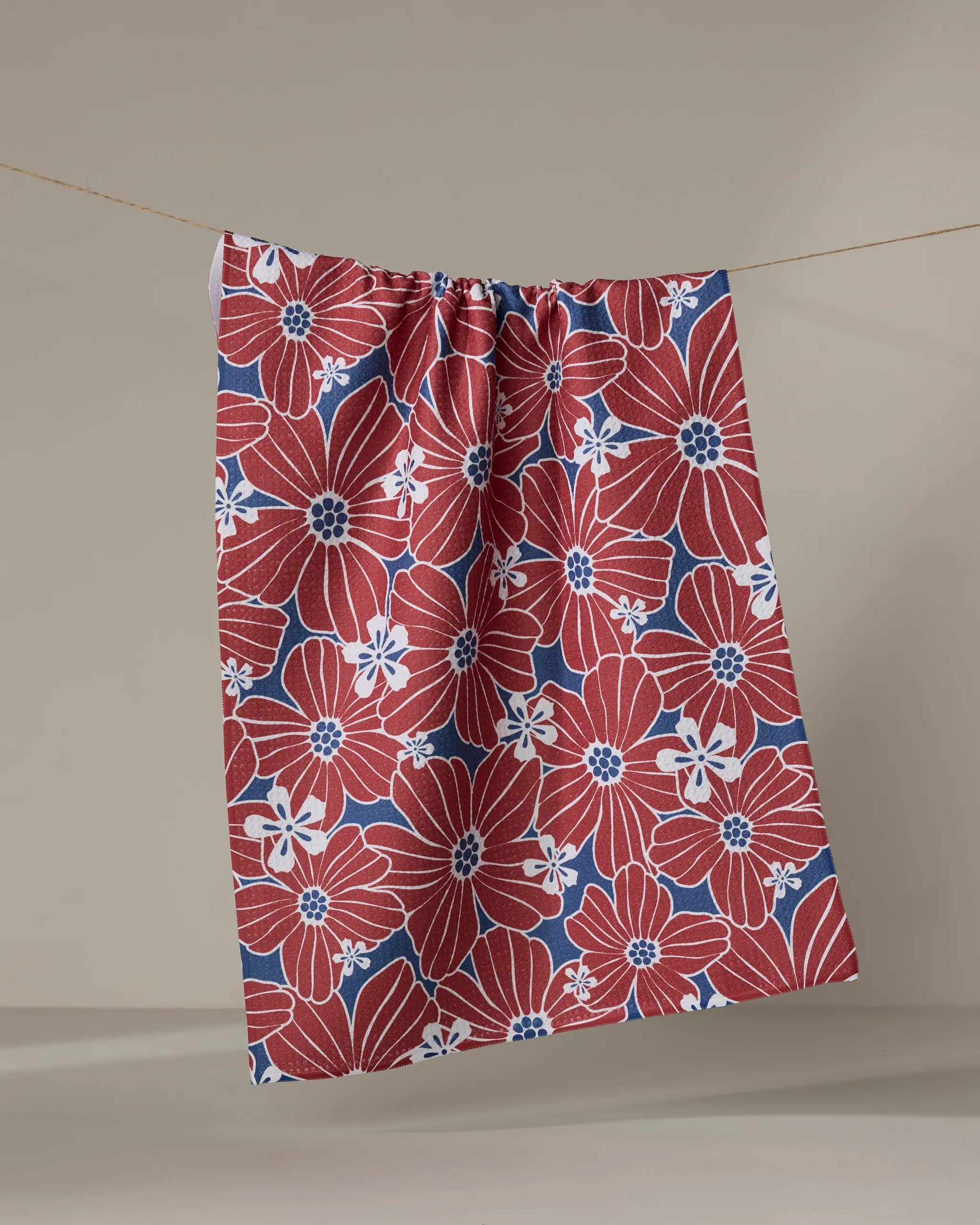 The Ashley Celena "Botanical Blush" tea towel, crafted from recycled materials, features large red flowers with white and blue accents in a floral pattern, draped over a clothesline against a neutral background.