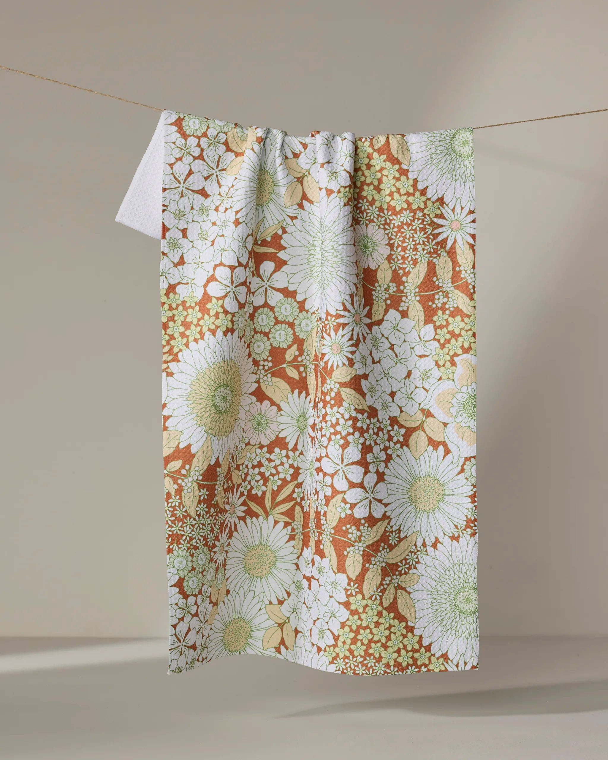 The Dabney Lee "Botanical Garden" recycled kitchen towel, featuring a vintage-inspired floral pattern in white, green, and soft orange, hangs on a clothesline against a neutral background.