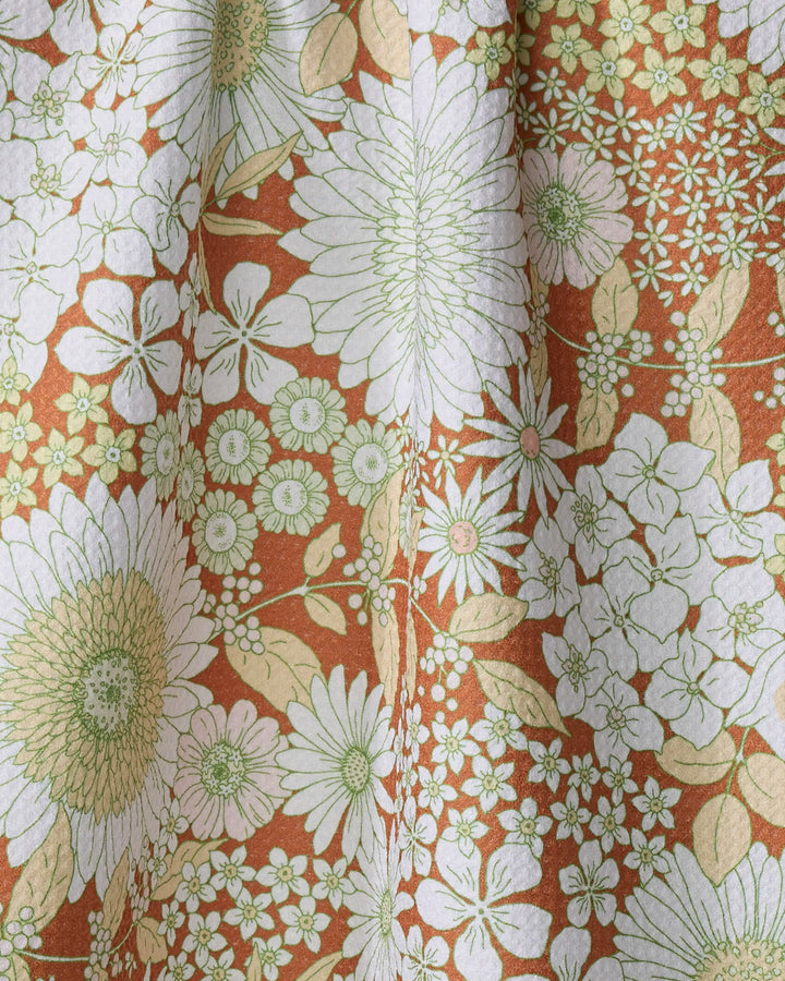 A close-up of the Dabney Lee "Botanical Garden" recycled kitchen towel, featuring a dense floral and leaf pattern in white, green, and beige on a rusty orange background.