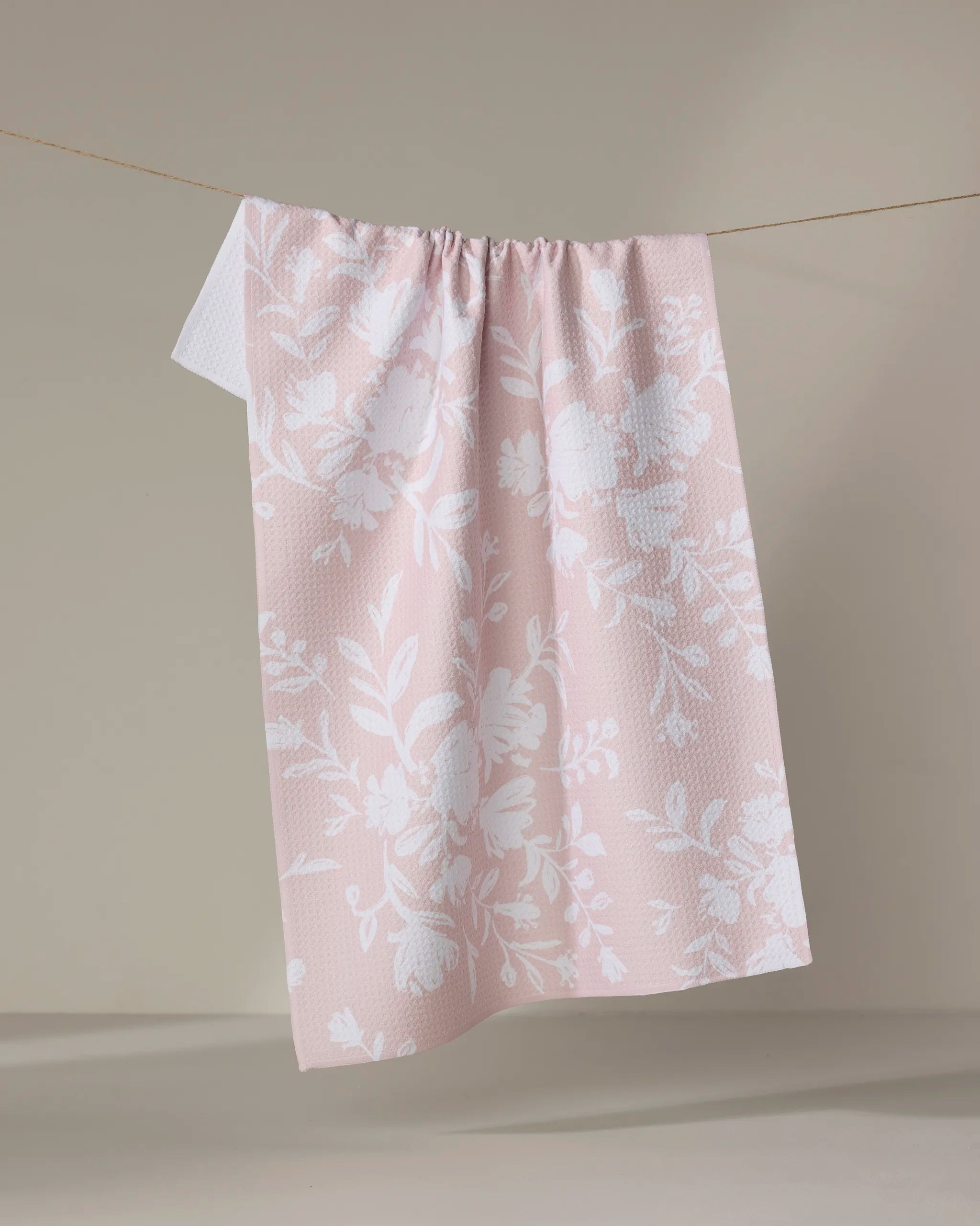 The Botanical Silhouettes Blush tea towel by Danelys Sidron, featuring a white floral pattern on pink and made from recycled materials, hangs neatly on a line set against a plain beige background.