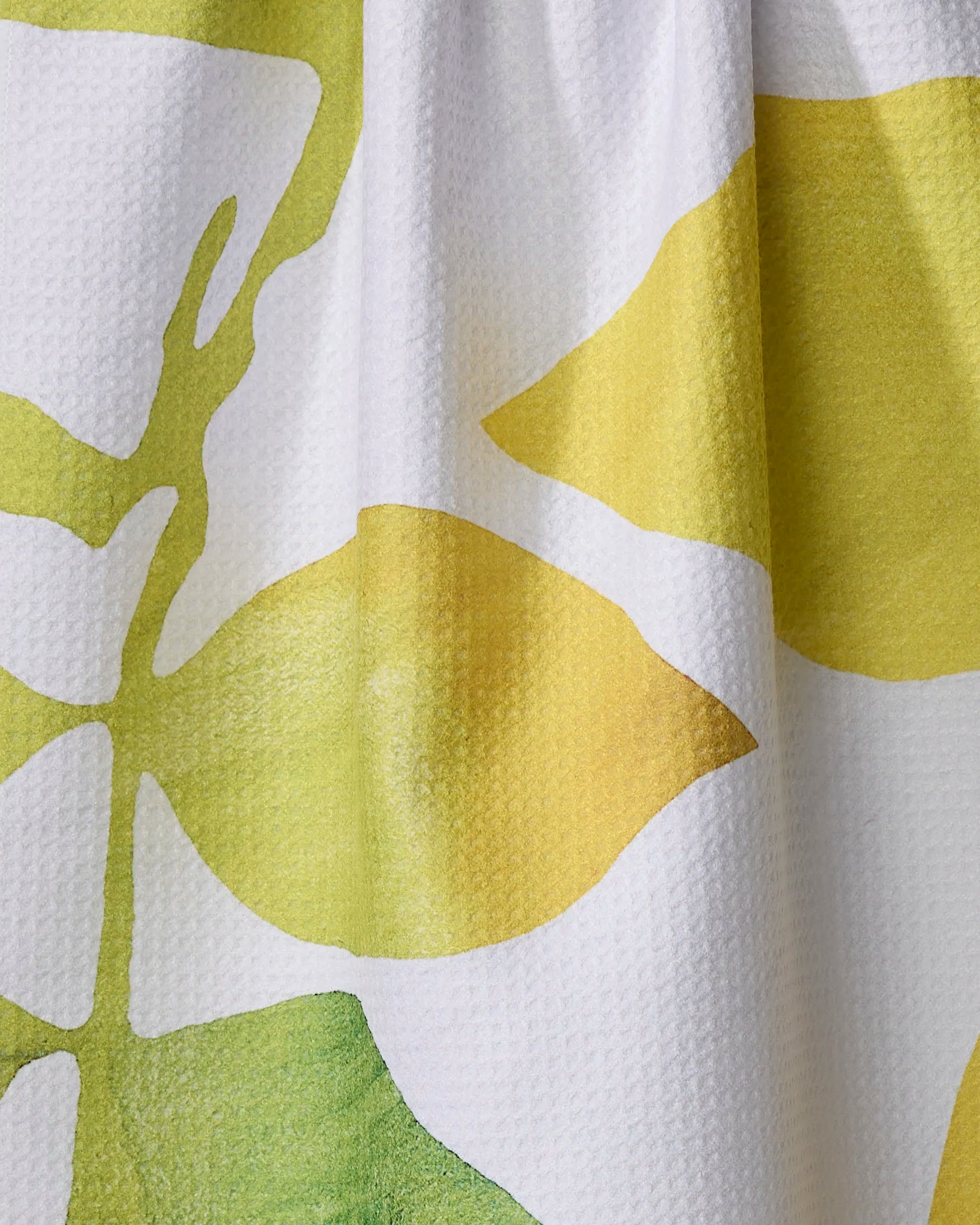 A close-up of Rachel Austin's "Botanical Watercolor Citrus" tea towel—white with a green and yellow abstract leaf pattern, crafted from recycled materials, showing its textured fabric, soft shadows, and gentle folds.