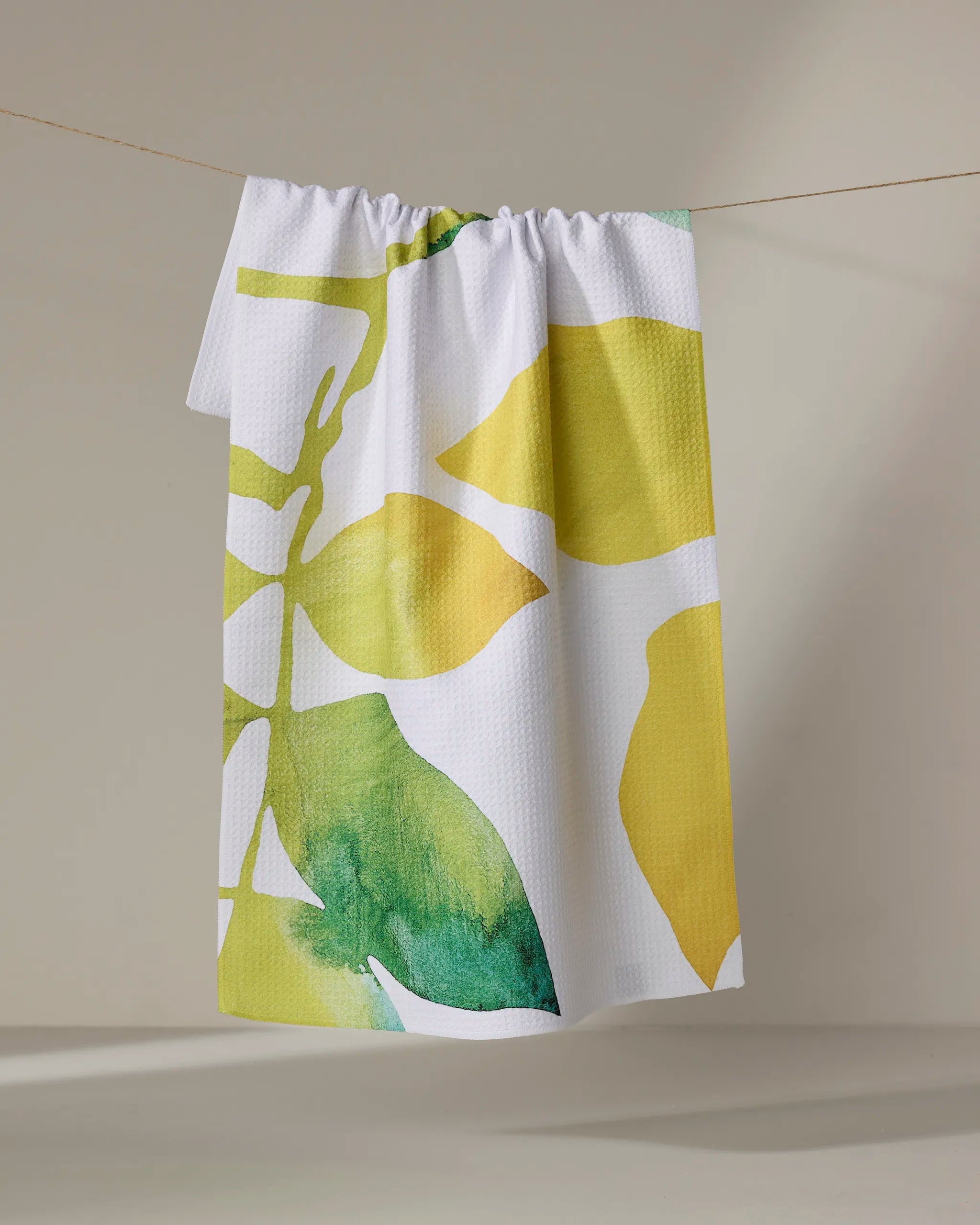 The "Botanical Watercolor Citrus" tea towel by Rachel Austin—crafted from ultra-absorbent recycled materials—features bold yellow and green abstract leaf patterns and hangs on a clothesline against a neutral background.