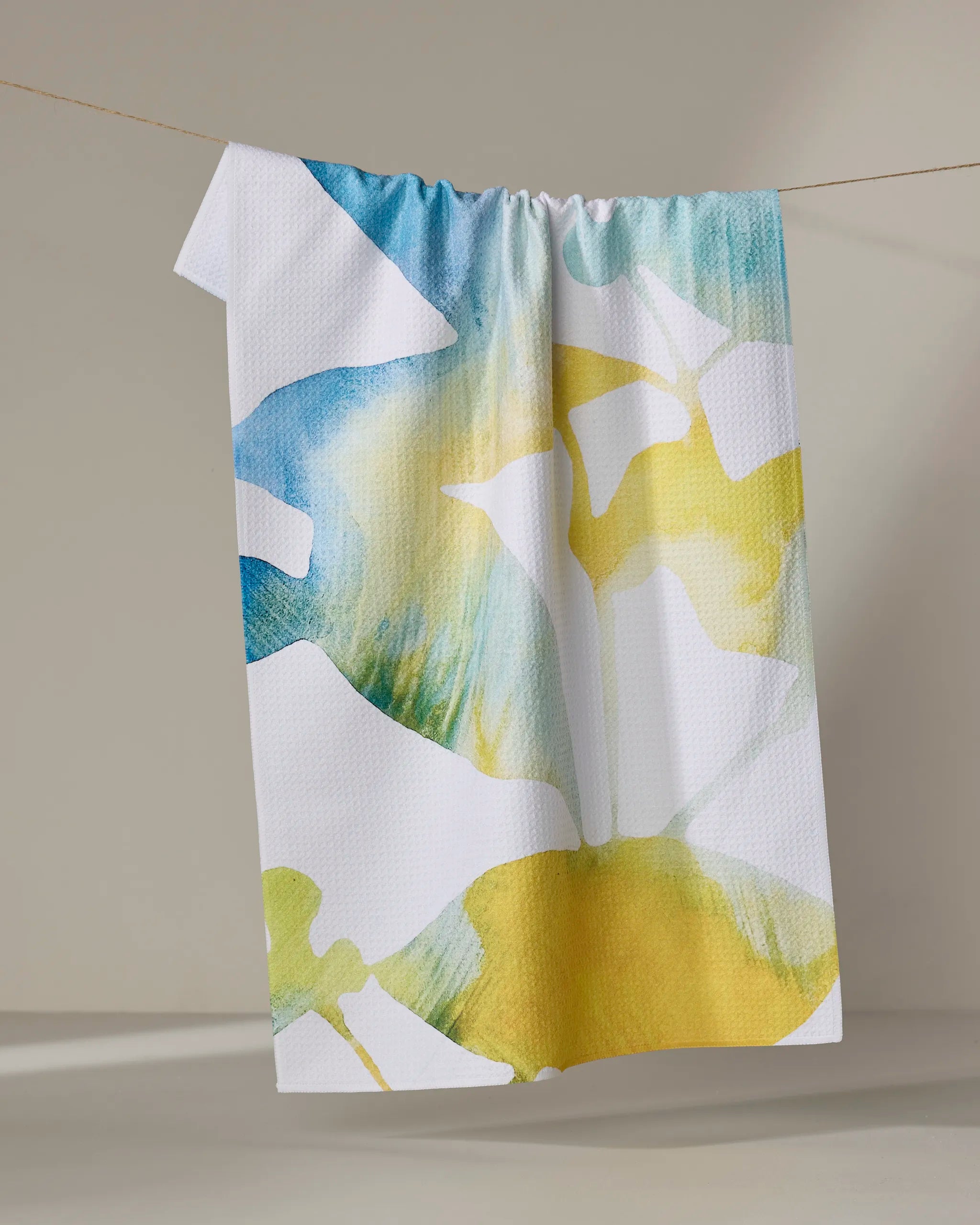 The "Botanical Watercolor Dews" tea towel by Rachel Austin, featuring ultra-absorbent fabric and abstract blue, green, and yellow patterns, hangs on a clothesline against a neutral beige background.