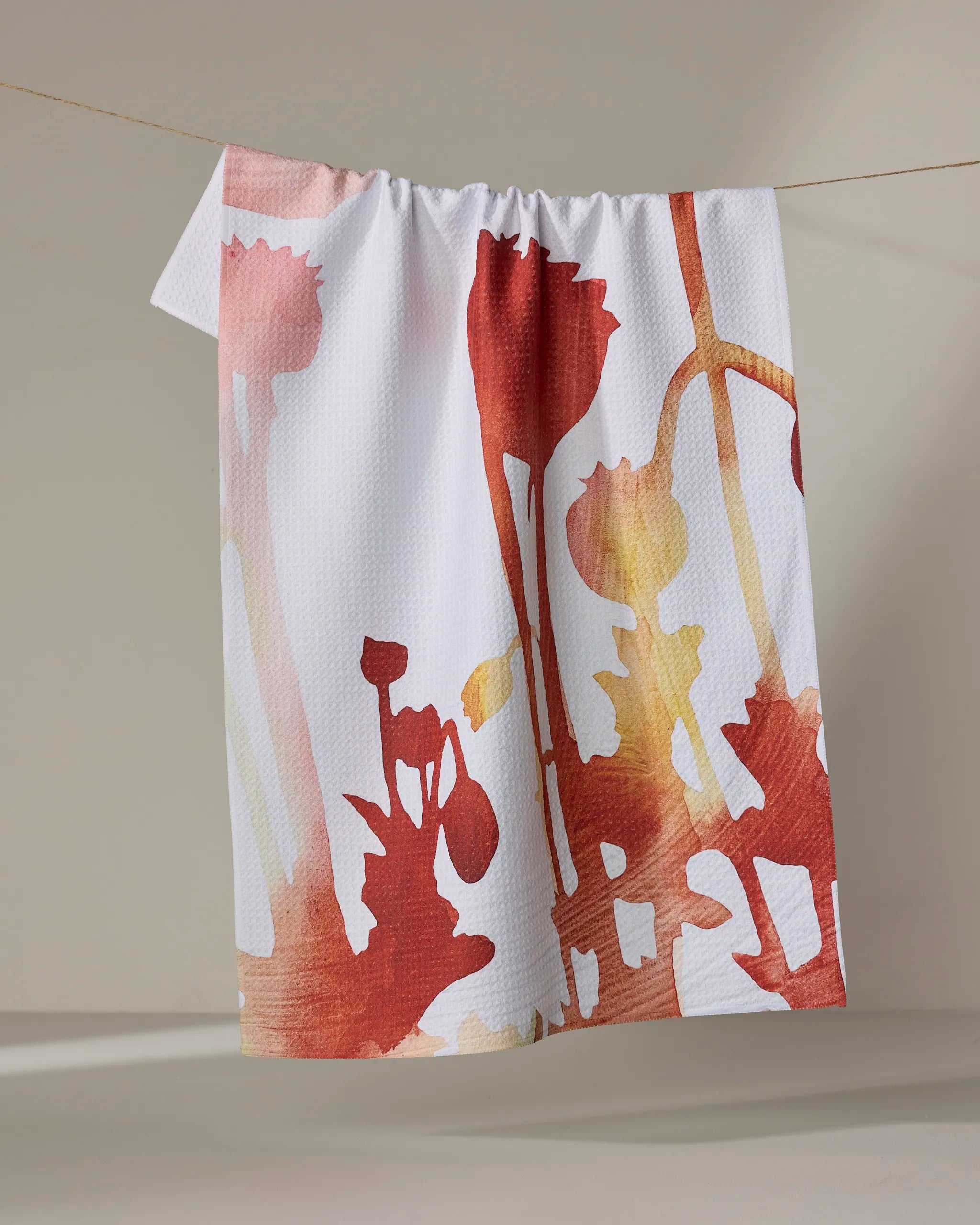 The Rachel Austin "Botanical Watercolor Dusk" towel, featuring a red and orange floral silhouette pattern on white, hangs on a clothesline against a neutral background.