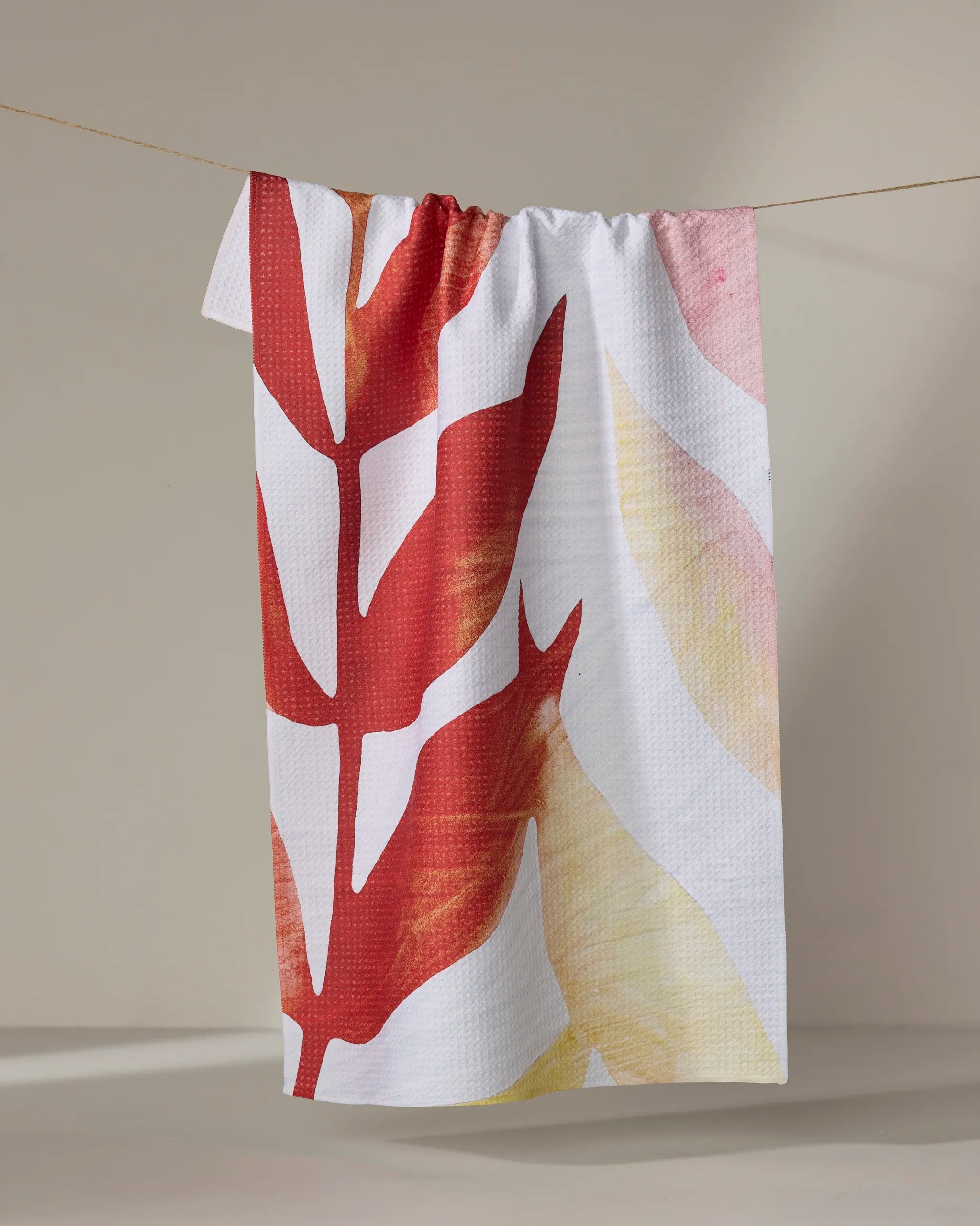 The "Botanical Watercolor Light" towel by Rachel Austin, featuring large red and yellow leaf patterns, hangs from a clothesline against a neutral background.