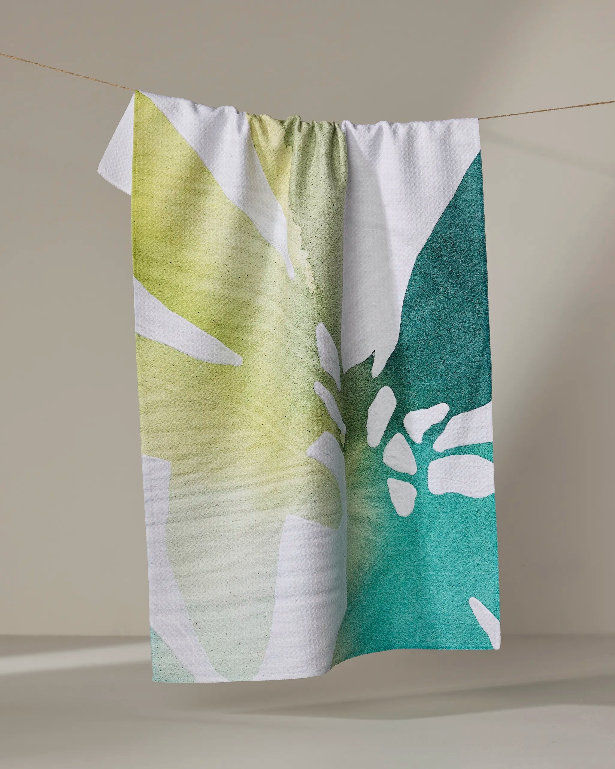 The "Botanical Watercolor Sea" quick-drying towel by Rachel Austin, featuring a large abstract leaf design in green, yellow, and teal, hangs on a clothesline against a neutral backdrop.