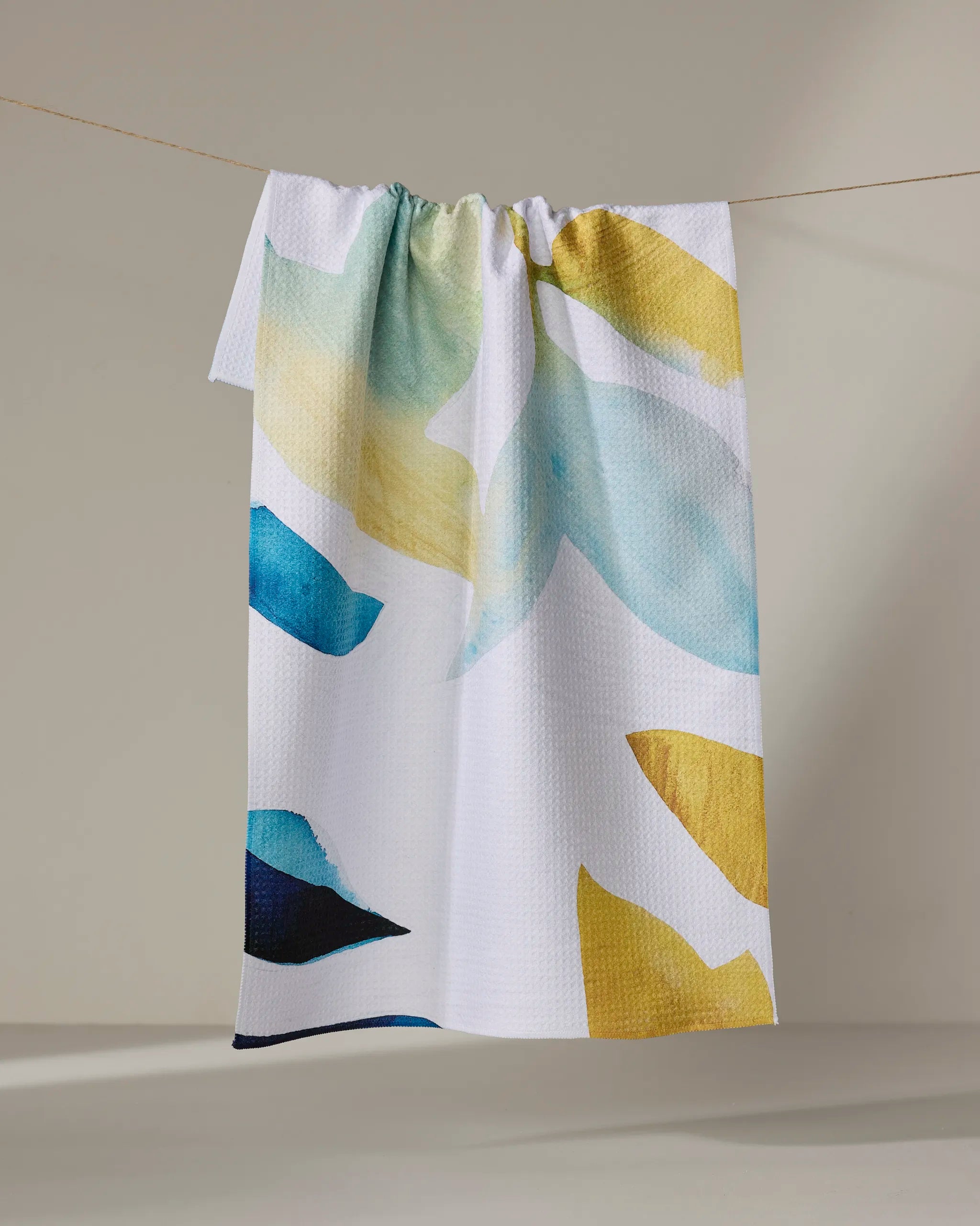 The Botanical Watercolor Spring tea towel by Rachel Austin features textured white fabric with abstract blue, yellow, and green shapes, made from recycled materials and shown hanging on a string against a neutral background.