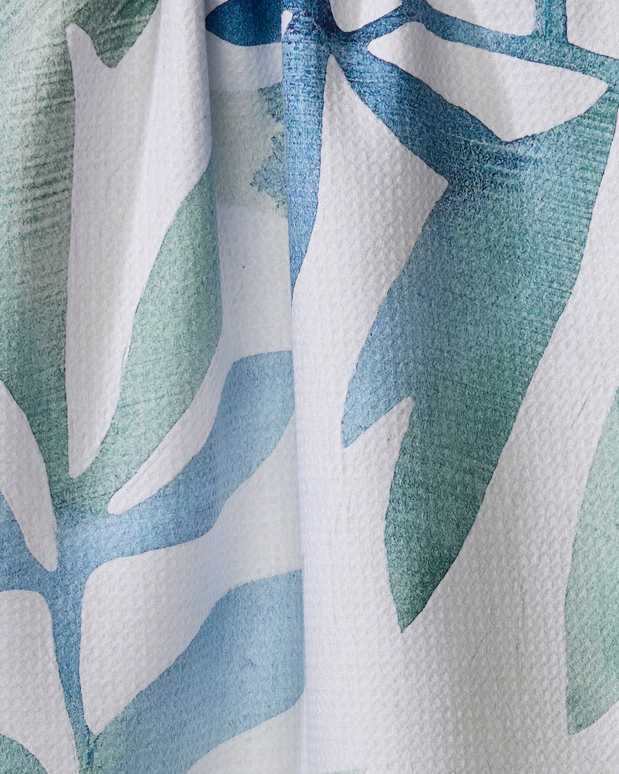 Close-up of Rachel Austin's Botanical Watercolor Tropic tea towel, showcasing an abstract blue and green leaf design on white. Made from recycled materials, the fabric displays visible folds and a slightly textured surface.