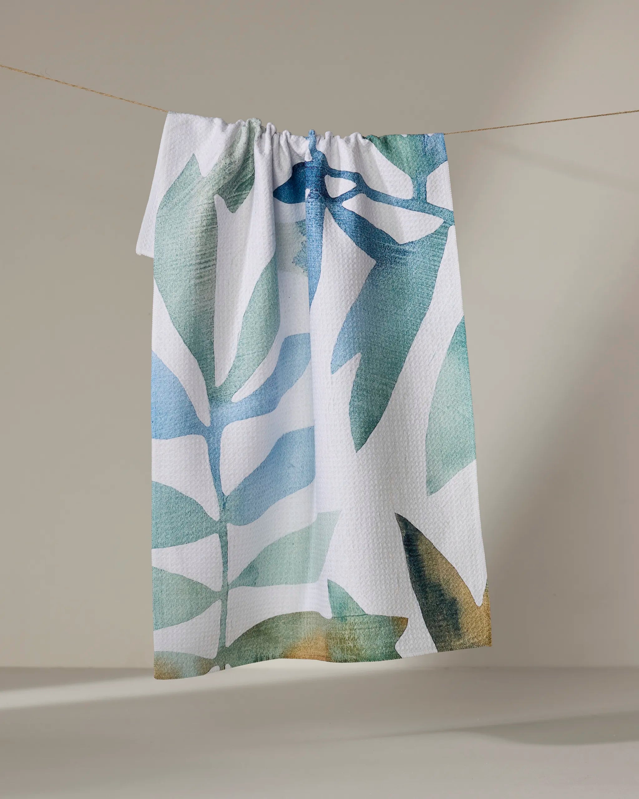 The Rachel Austin Botanical Watercolor Tropic tea towel, made from recycled materials, features large abstract green and blue leaf patterns. It hangs on a thin clothesline against a plain background and is both eco-friendly and quick-drying.